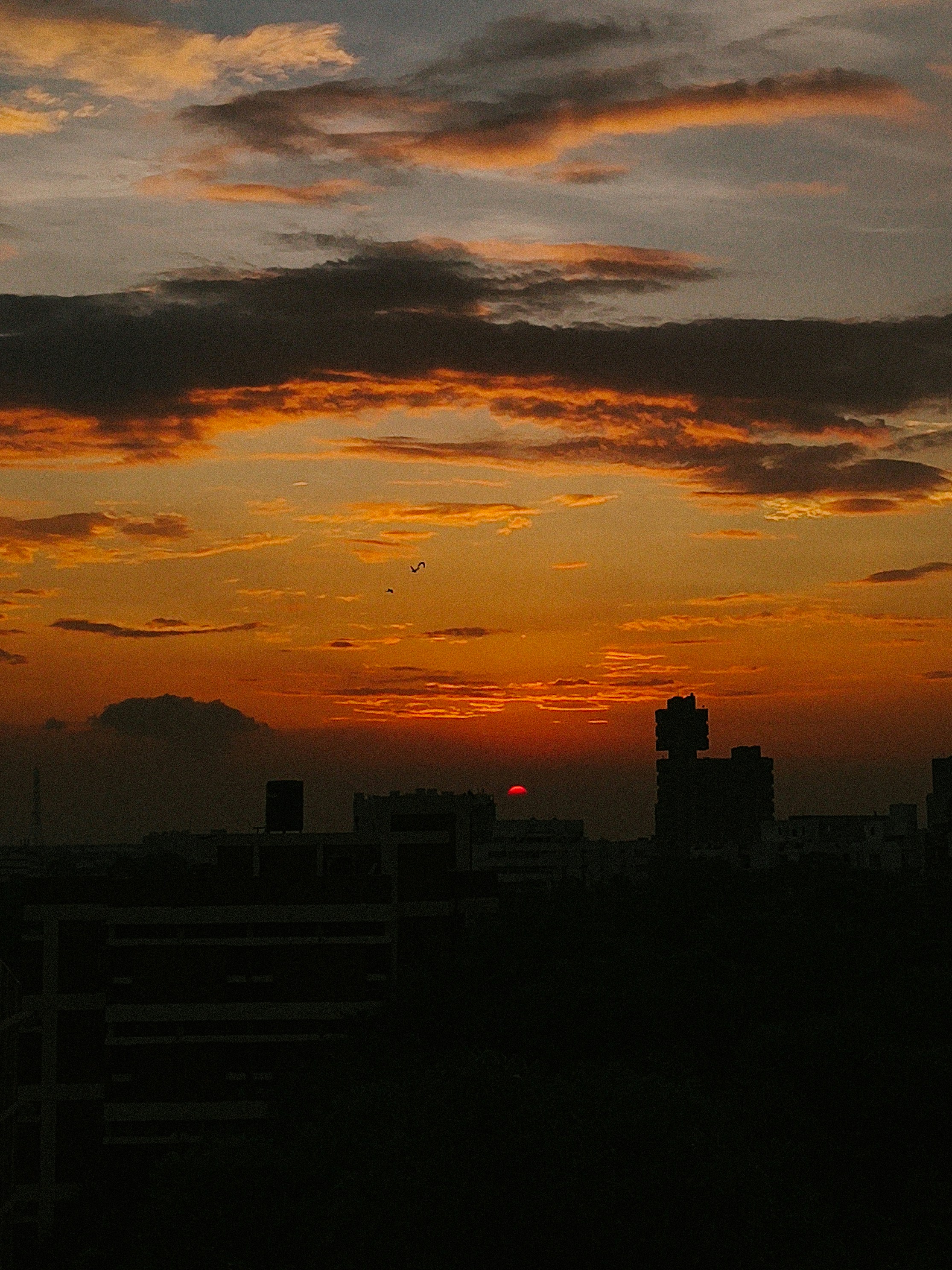 Un soleil bas brillant derrière des silhouettes sombres de bâtiments urbains sous un ciel orange chaud
