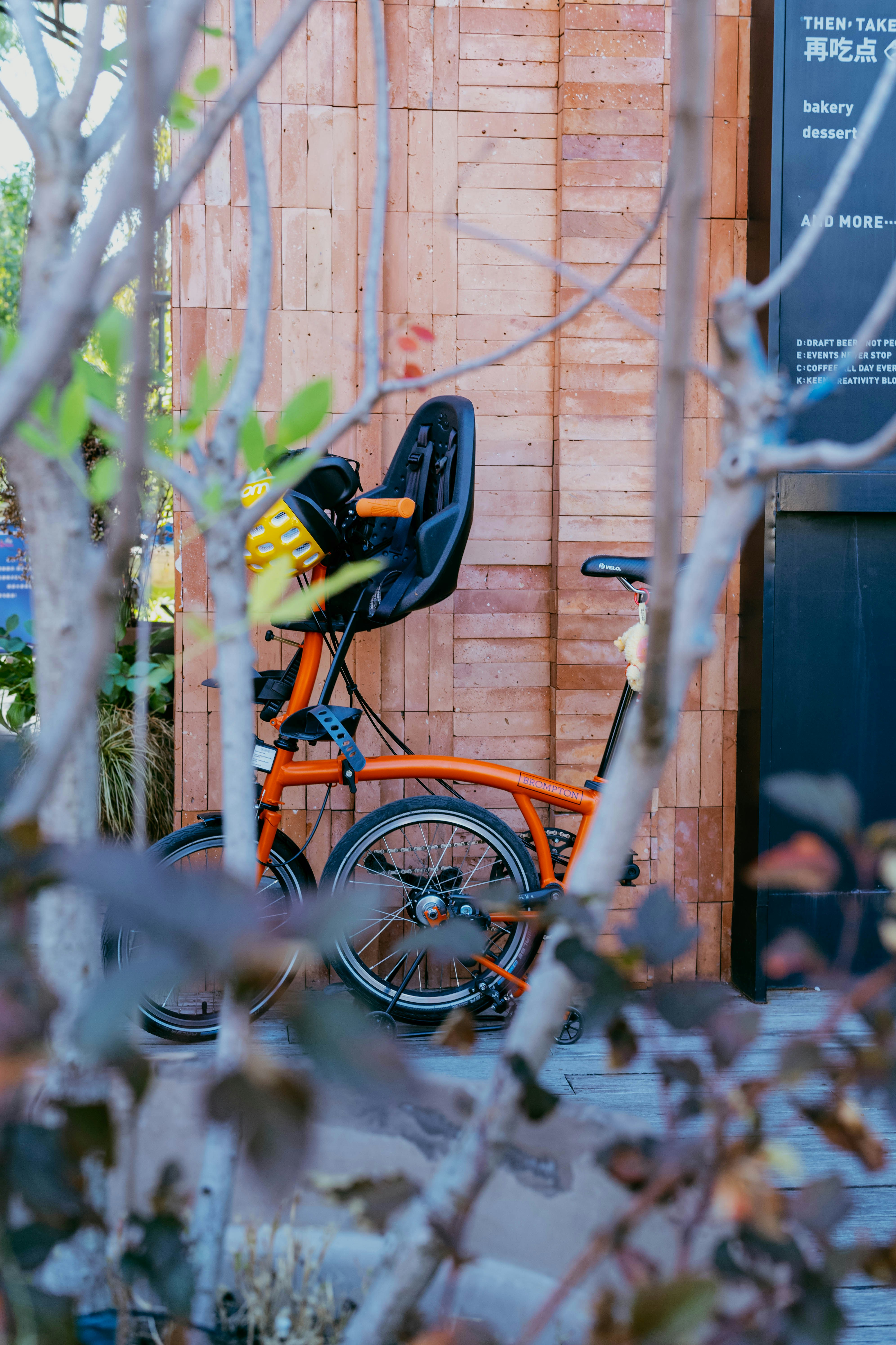 An orange folding bicycle with a child seat against a brick wall