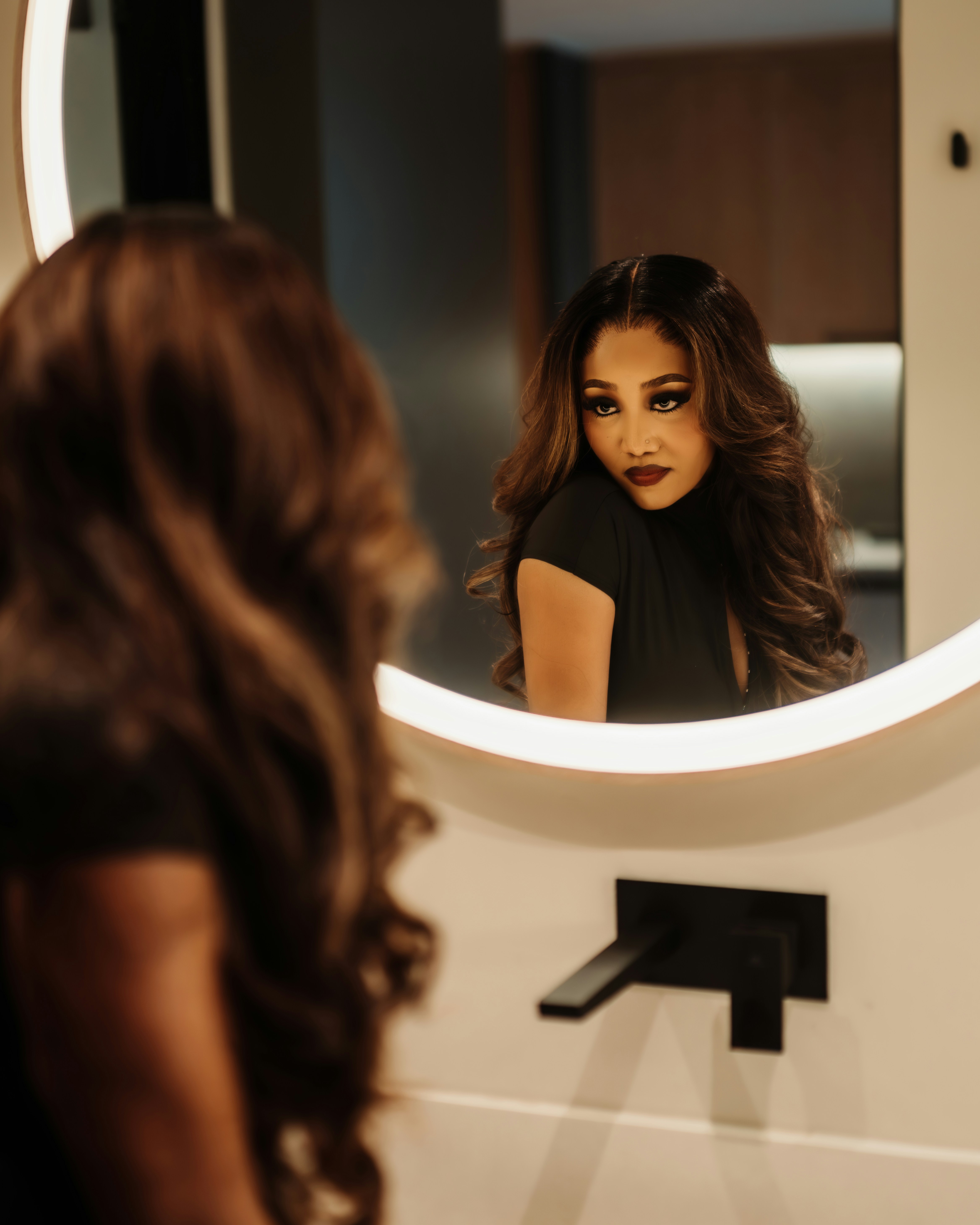 A woman with long wavy hair looking into a round illuminated bathroom mirror