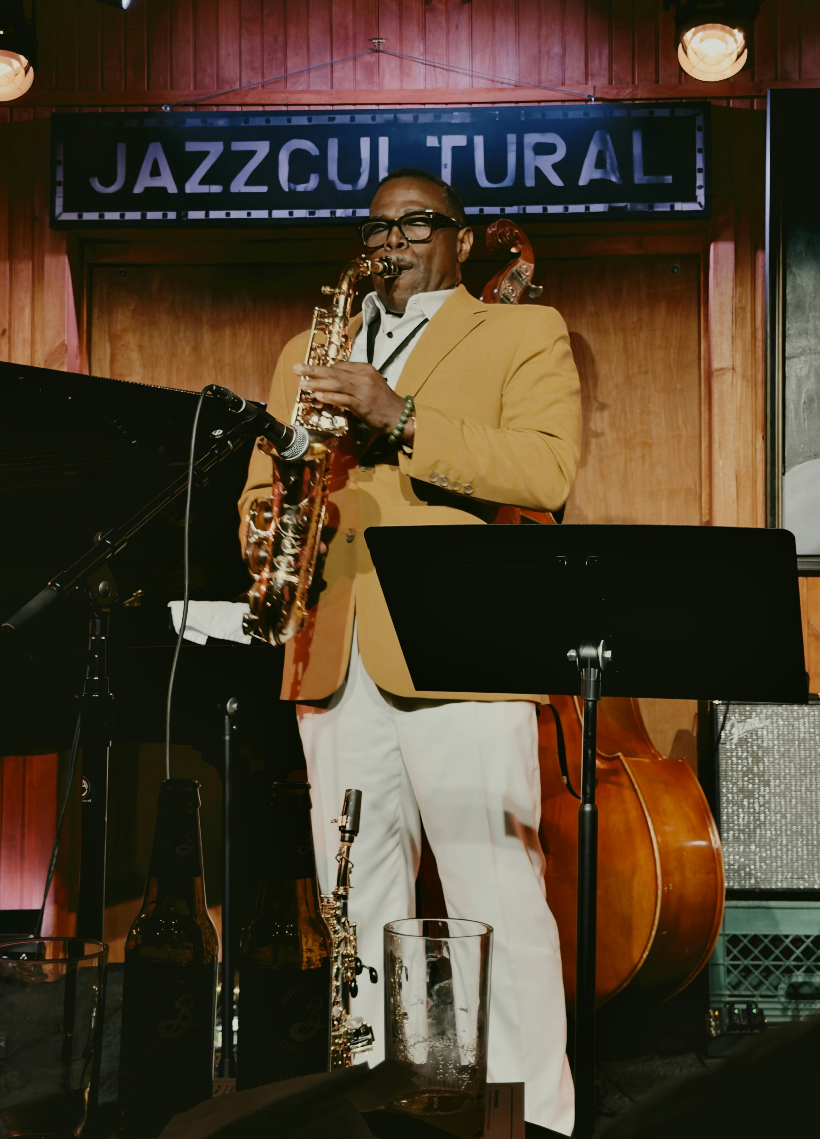 A man in a mustard blazer playing saxophone on a stage with a double bass