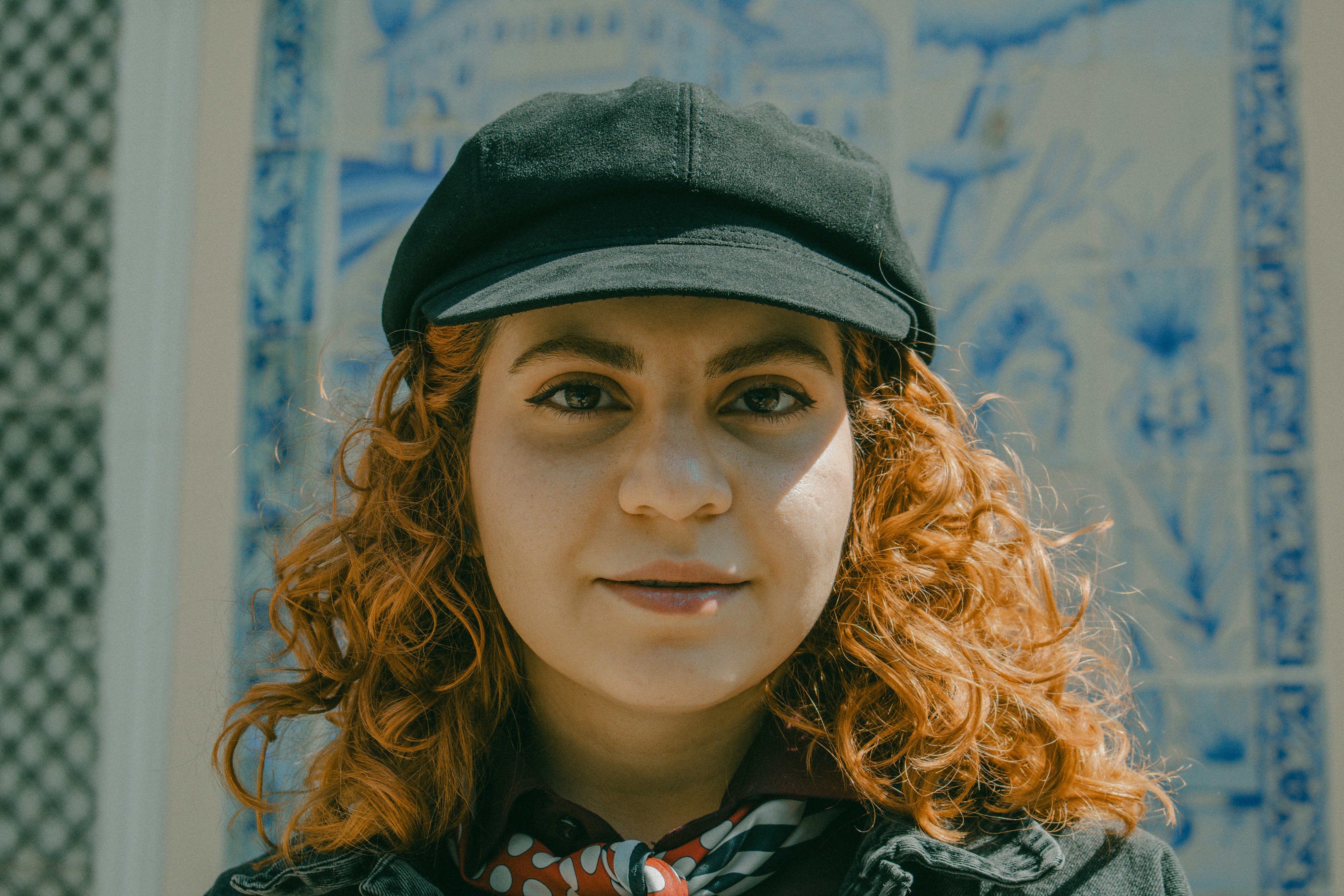 A woman with curly red hair wearing a black cap and a patterned scarf