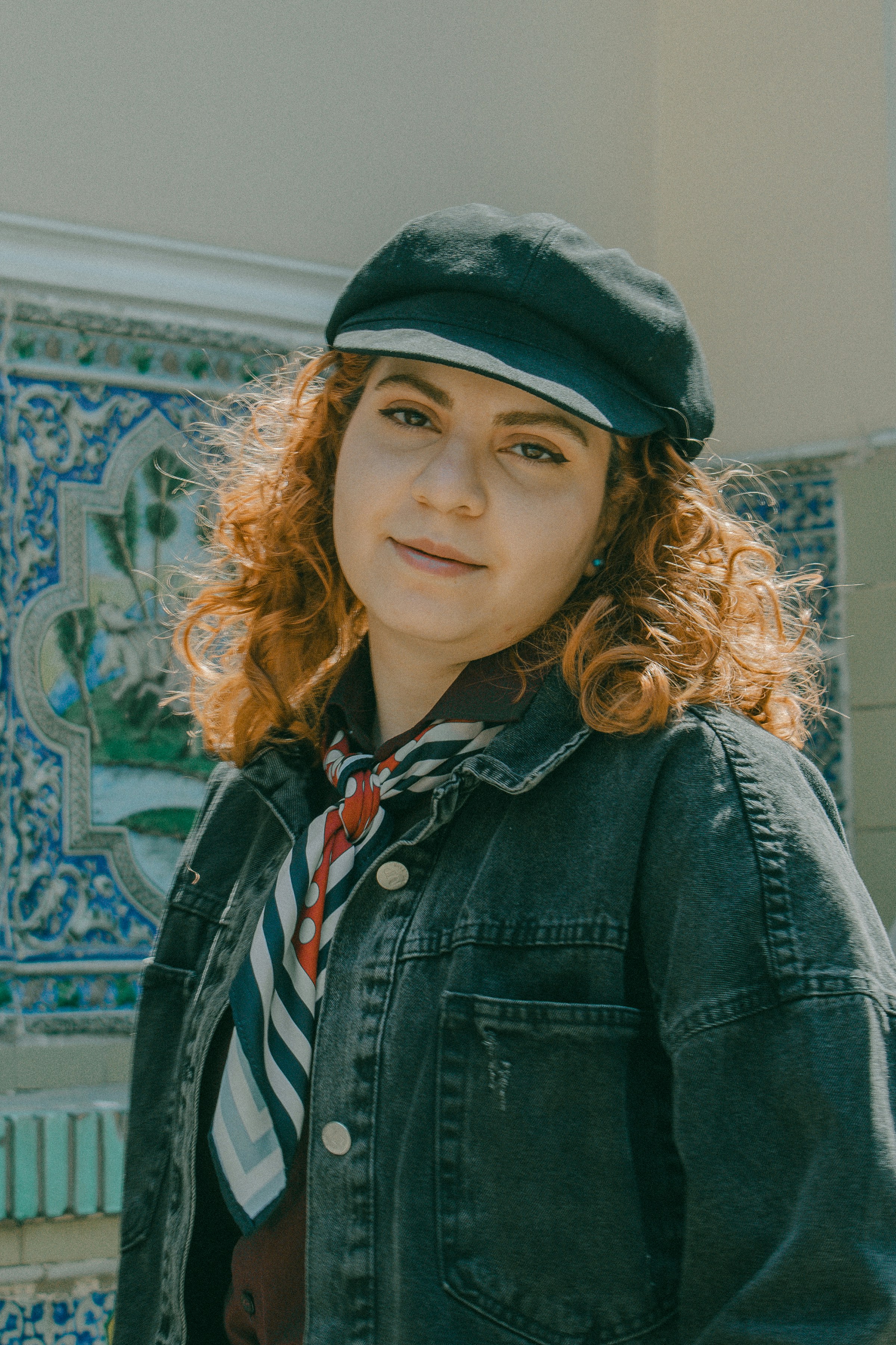 A woman with curly red hair wearing a black cap and denim jacket