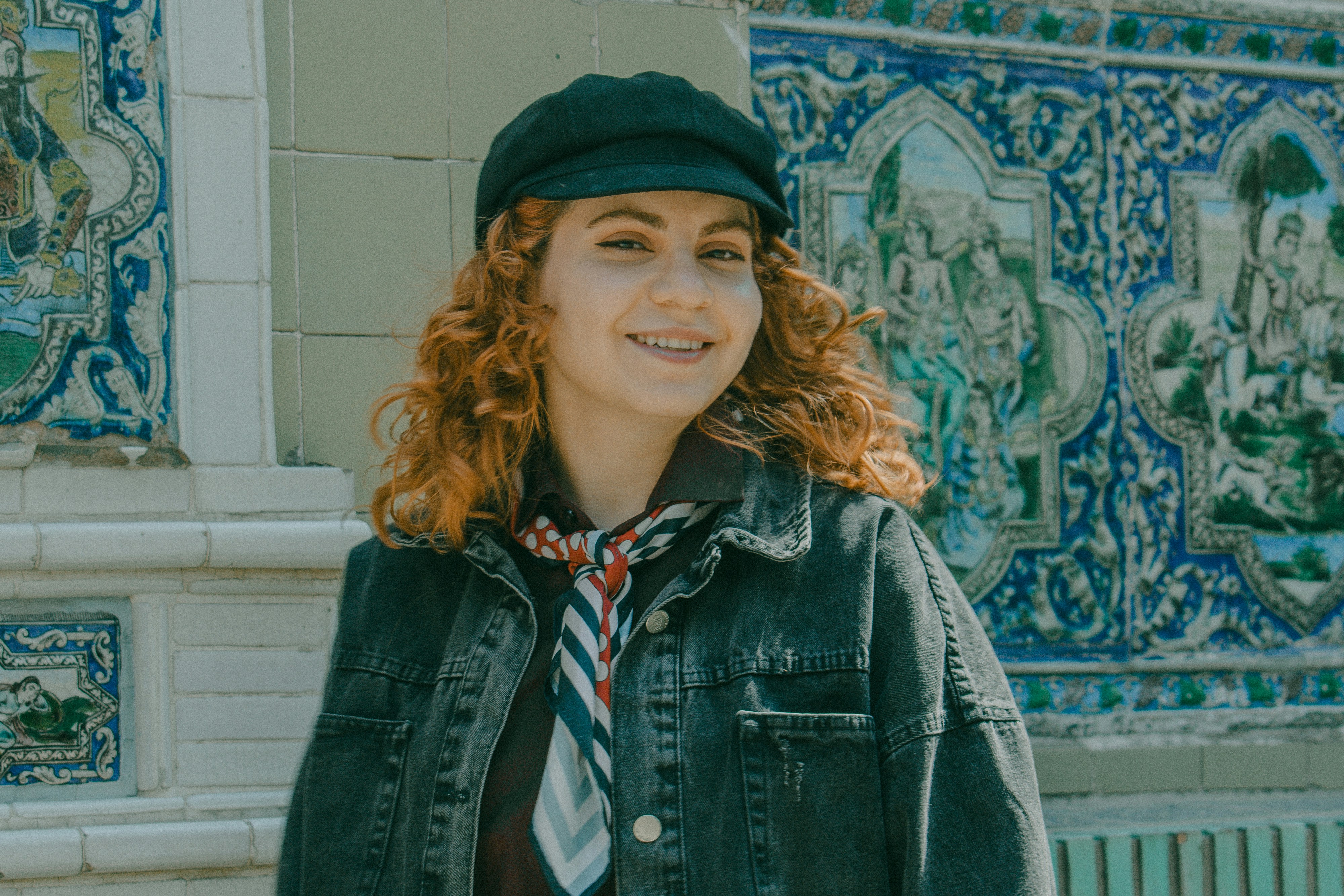 A woman with curly red hair wearing a black cap and denim jacket