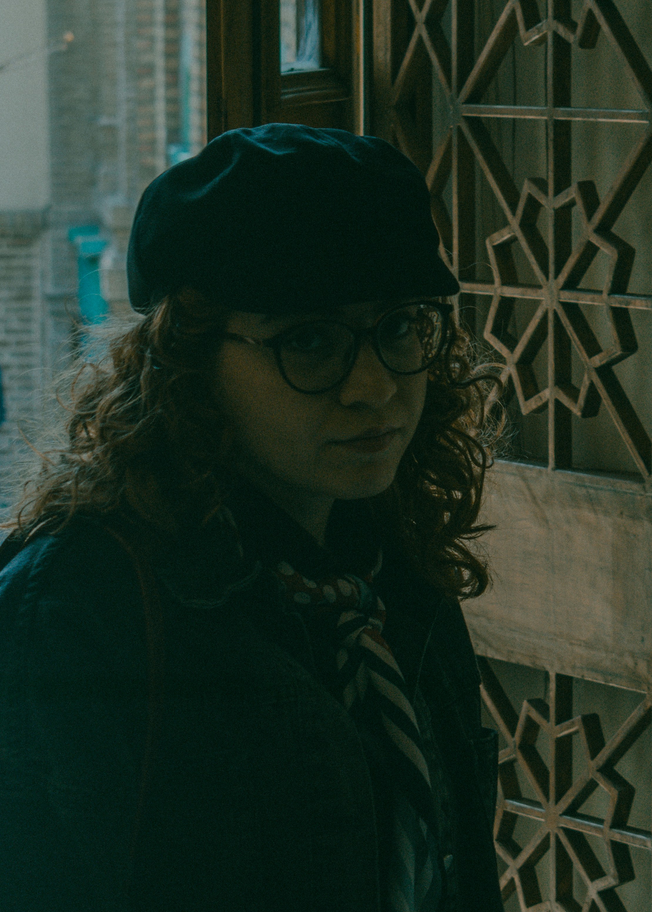 A person with curly hair wearing a cap and glasses by a patterned door