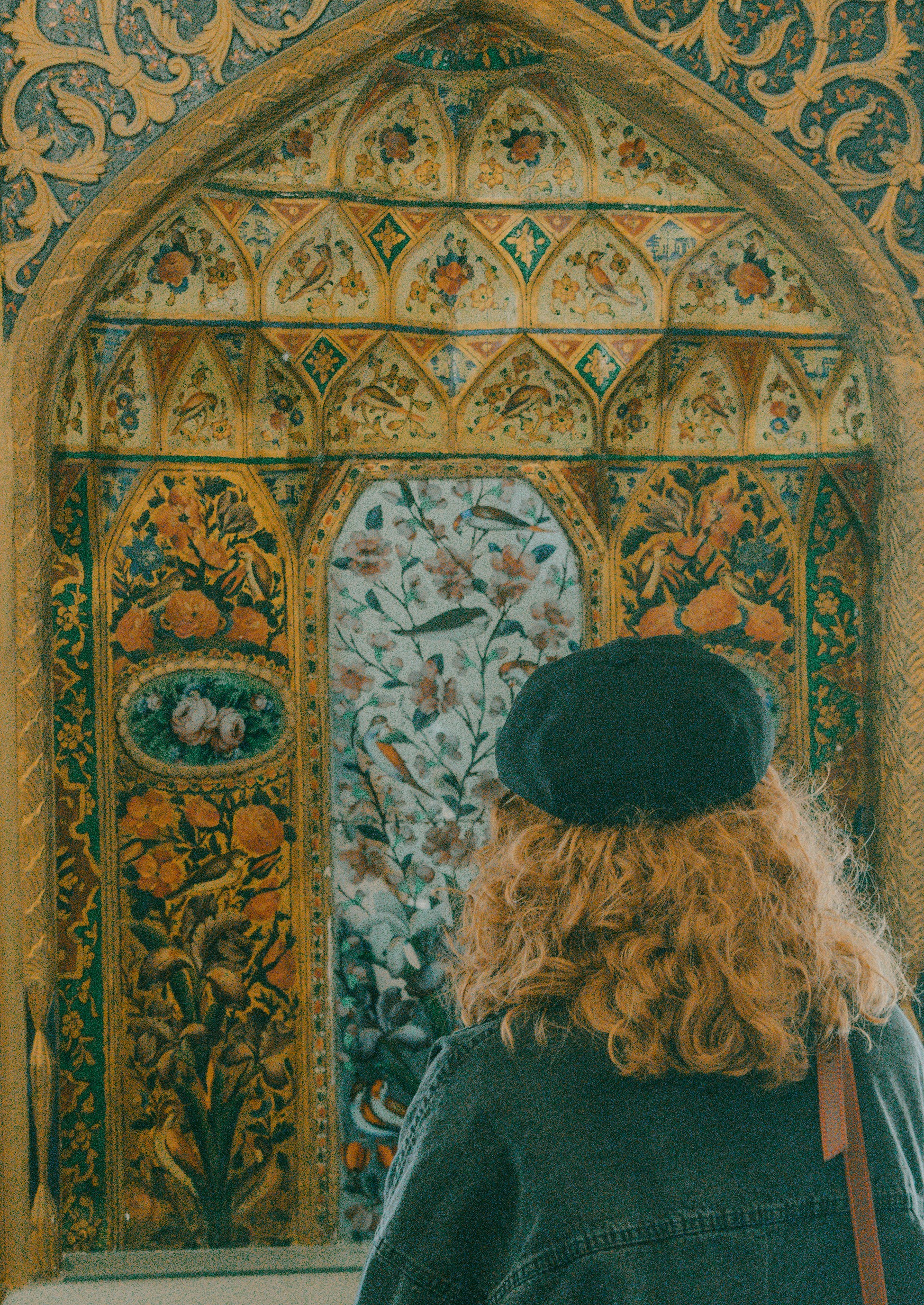 A person with curly hair and a beret viewing an ornate floral wall mural