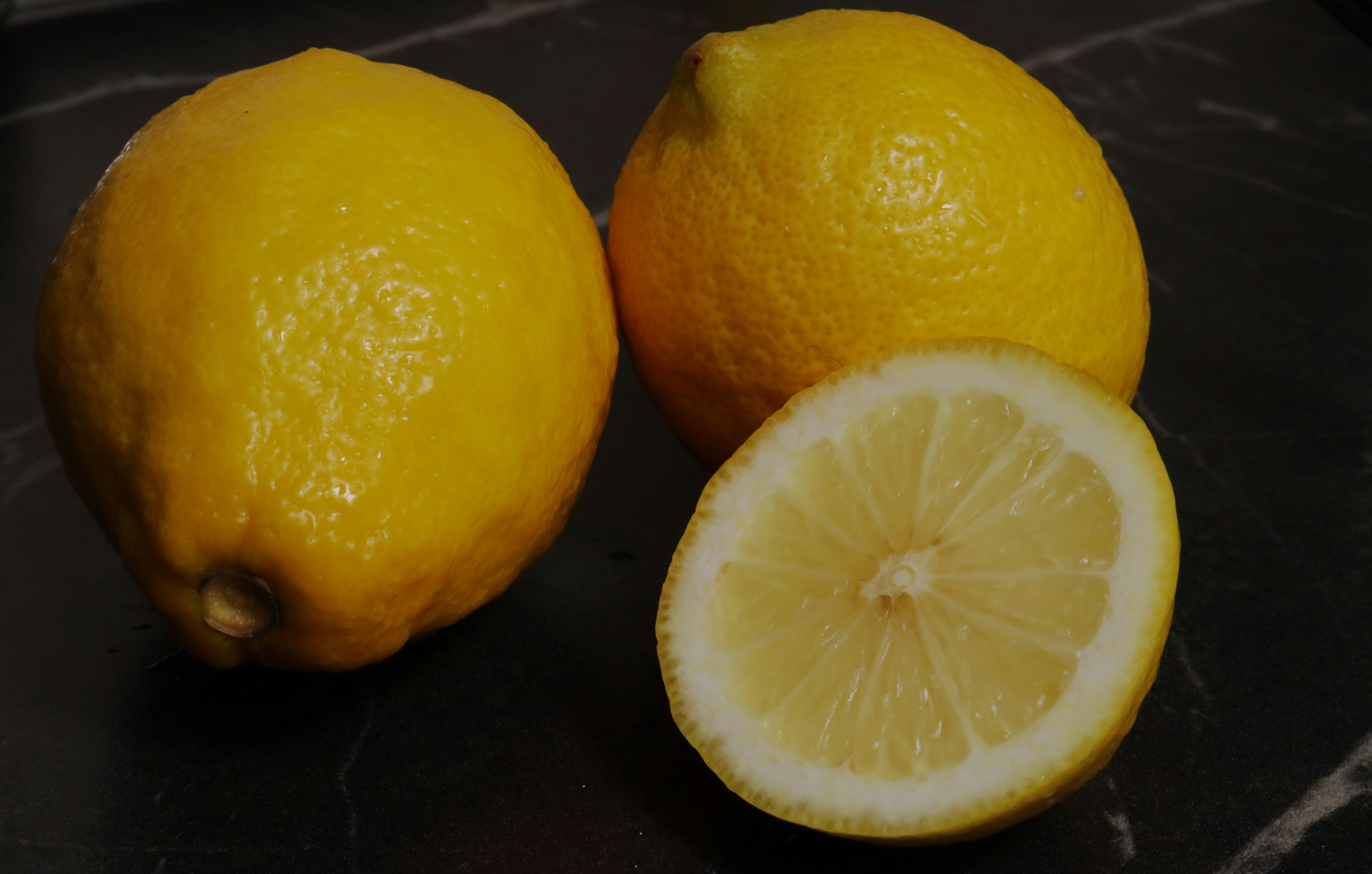 Two whole yellow lemons and one halved lemon on a dark surface