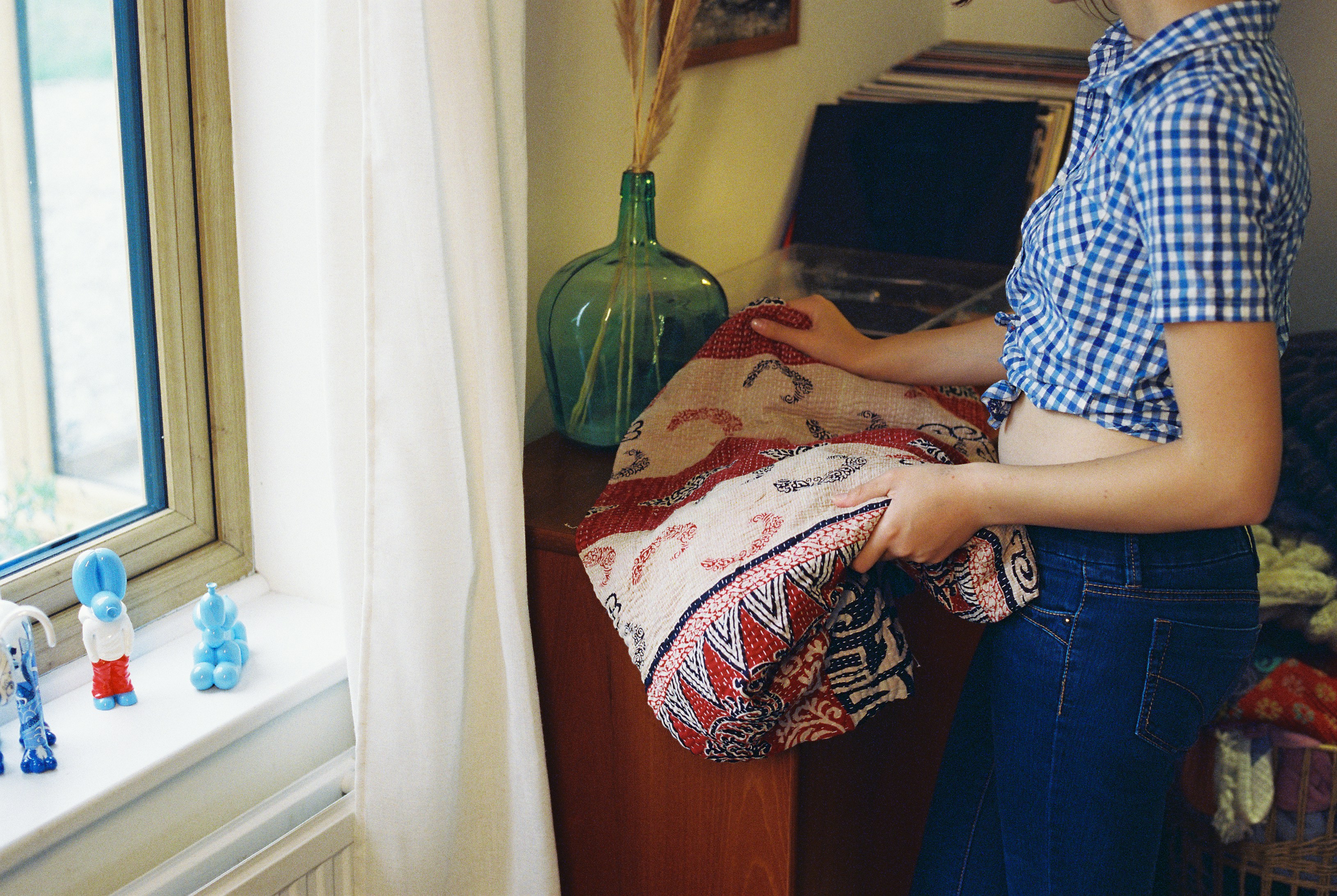 A woman holding a patterned quilt beside a teak sideboard and sunlit window