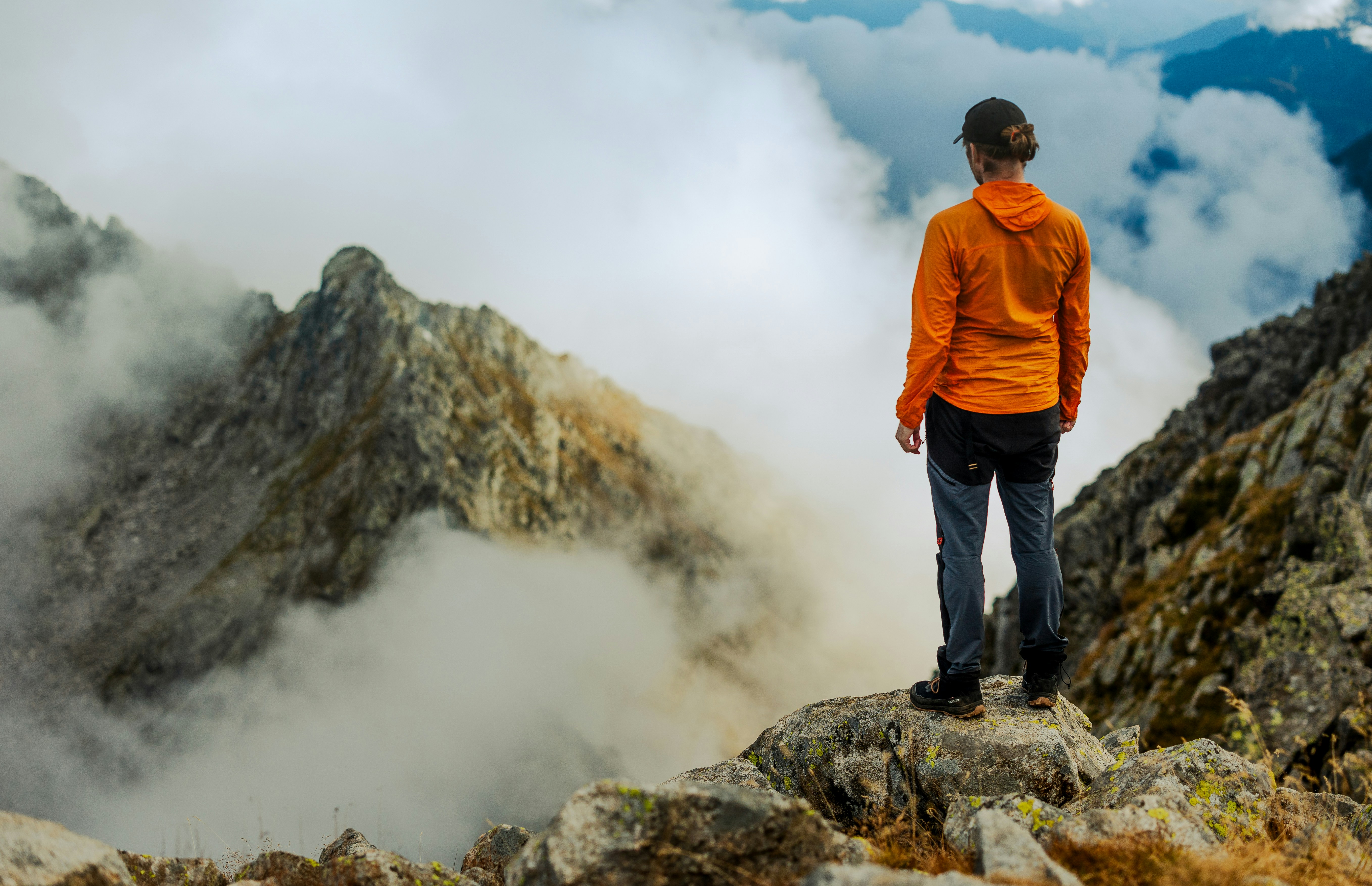 A person in an orange jacket on a rocky mountain peak above clouds