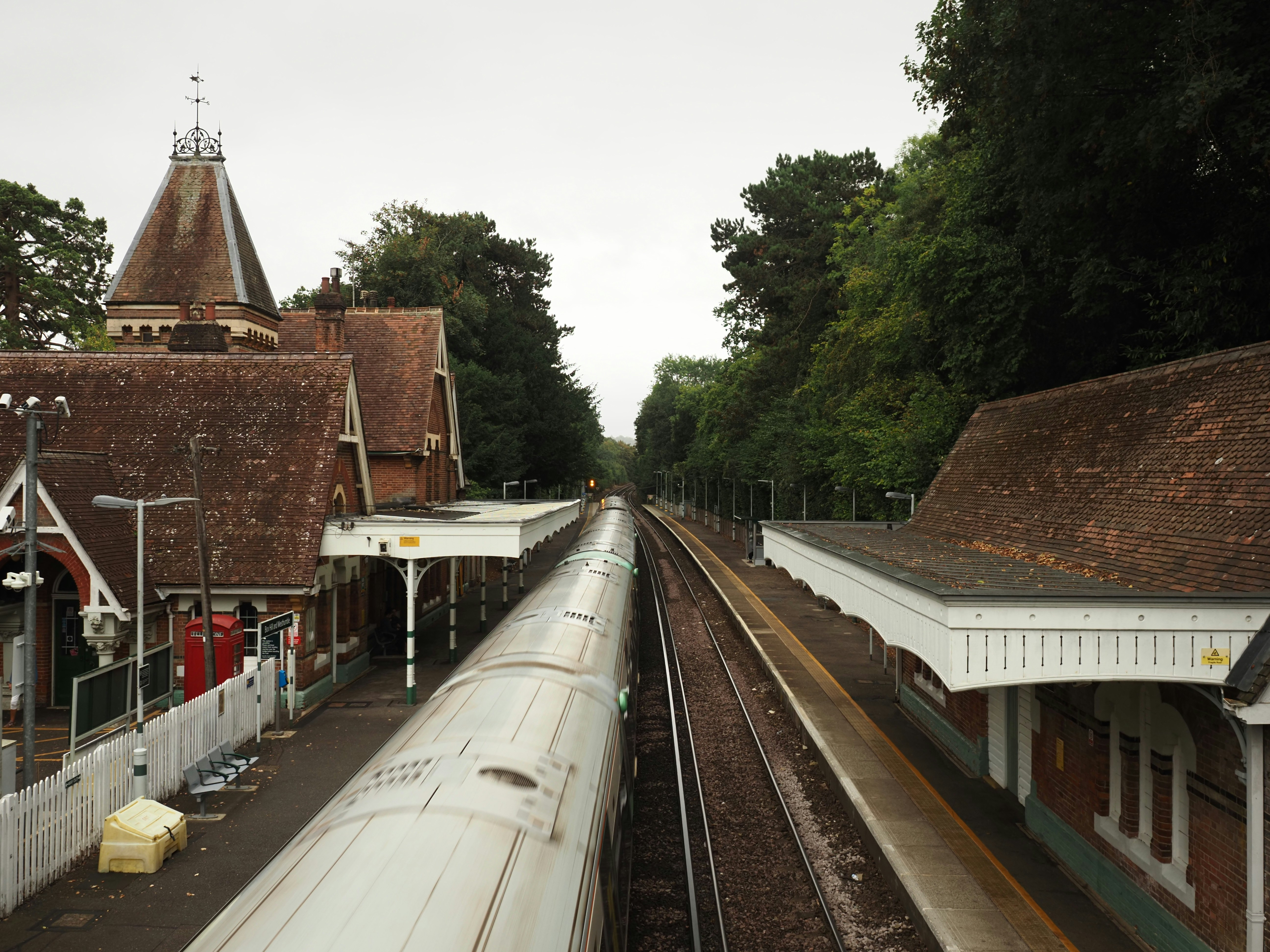 A train passes through a traditional English railway station, framed by Victorian architecture, tiled roofs and surrounding woodland.
