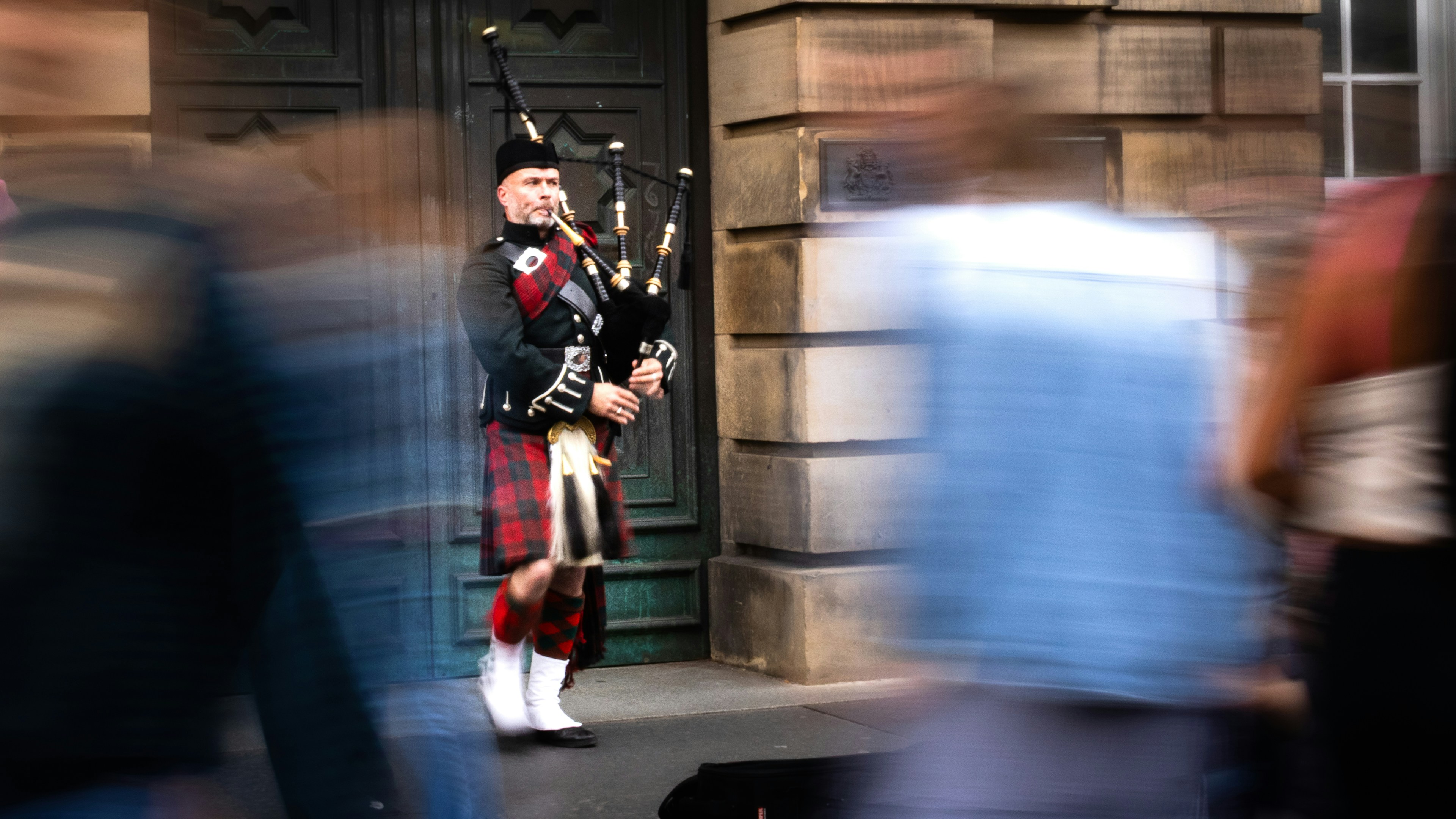 A bagpiper in traditional tartan kilt playing music before a blurred crowd