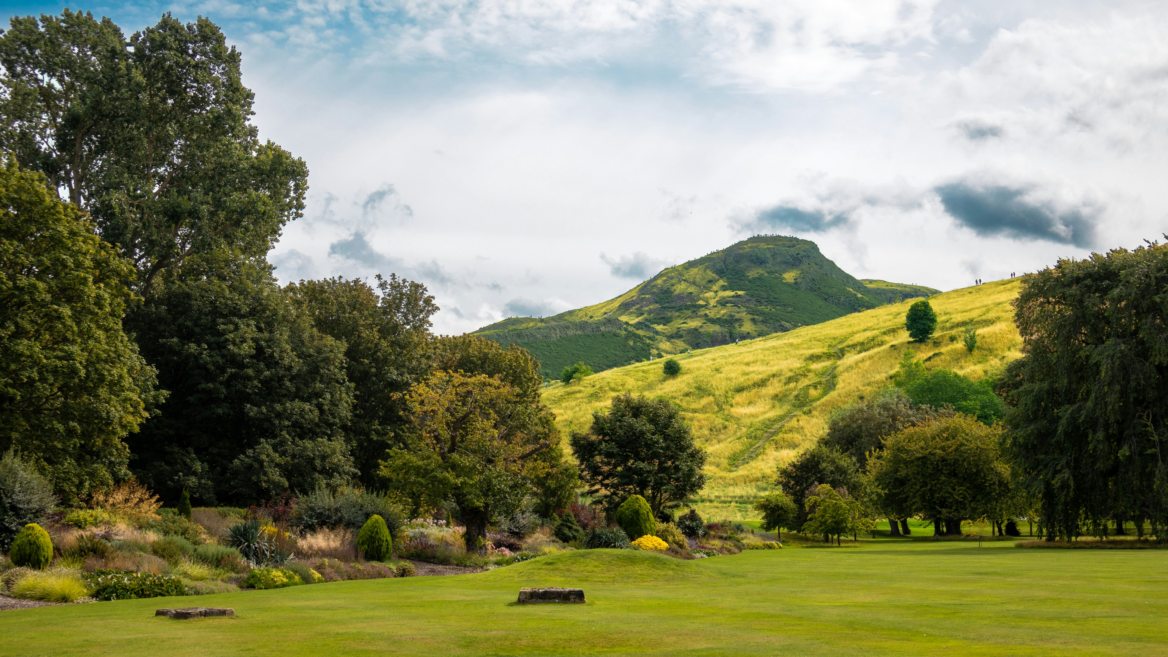 A green park with trees and shrubs below a grassy hill under clouds