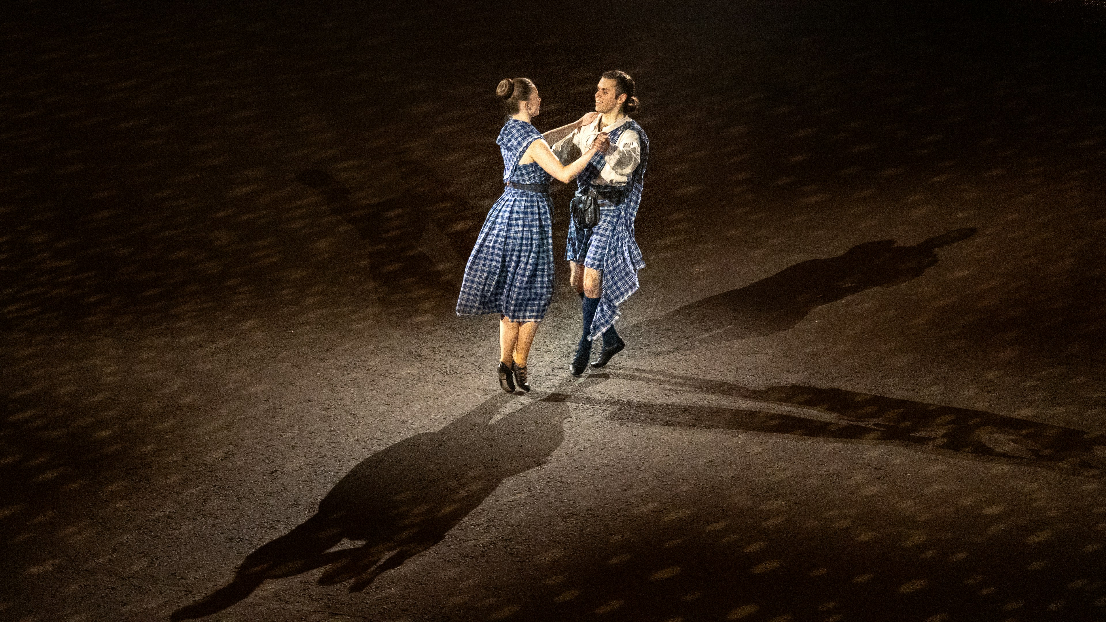 Two people in blue tartan kilts dancing on a dark stage with spotlights