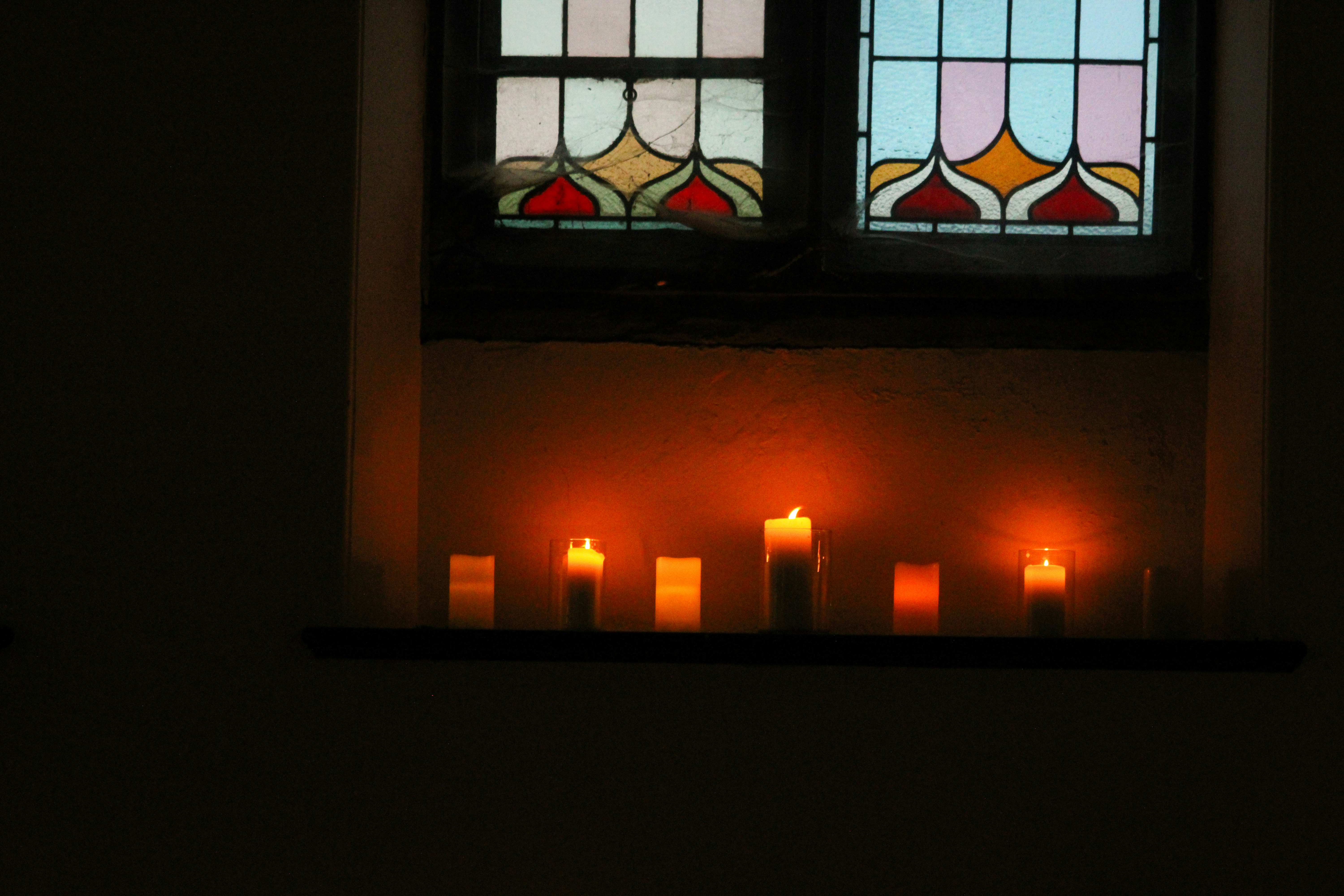 Lit candles on a ledge below a stained glass window in a dark room