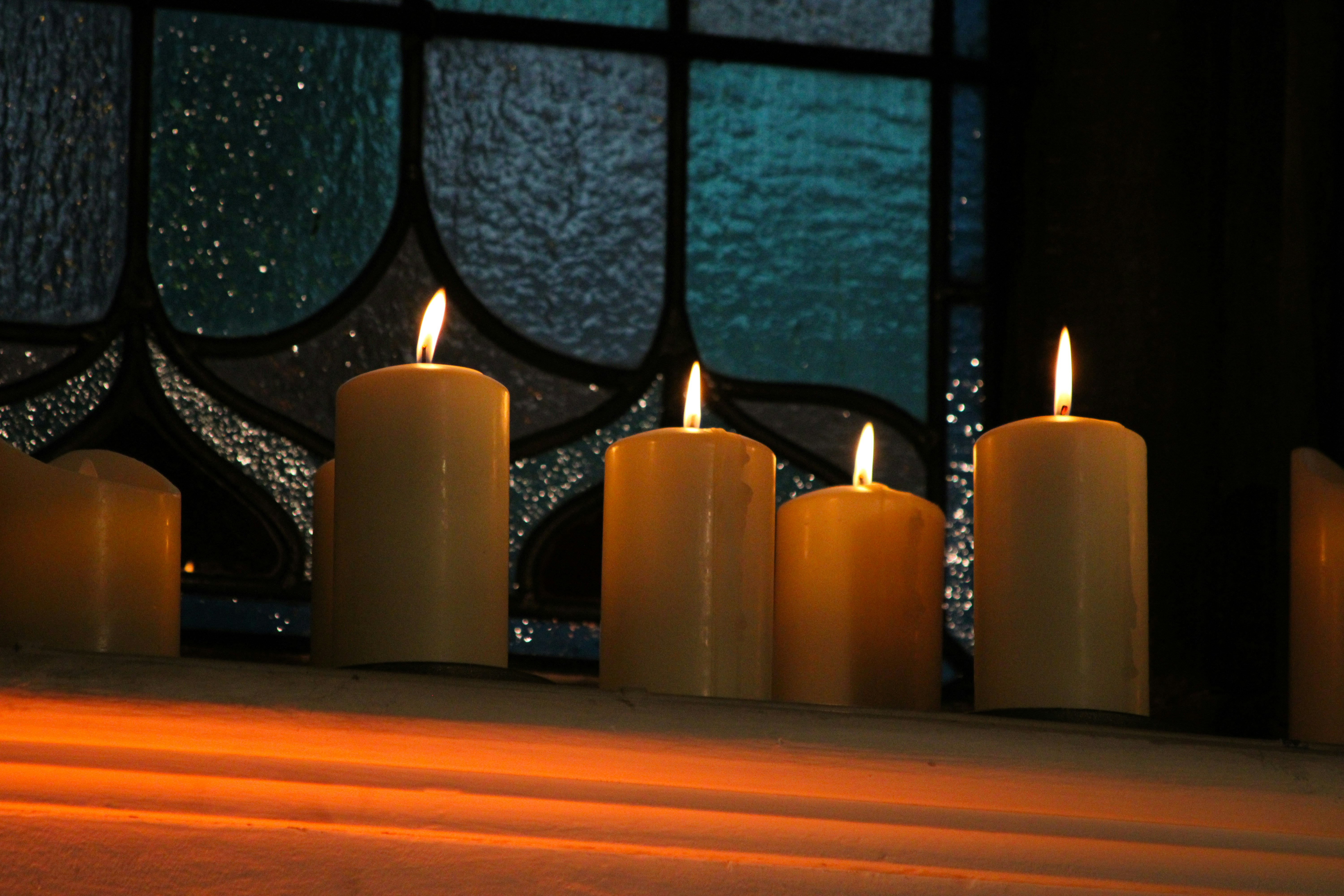 Four lit pillar candles on a ledge before a blue stained glass window