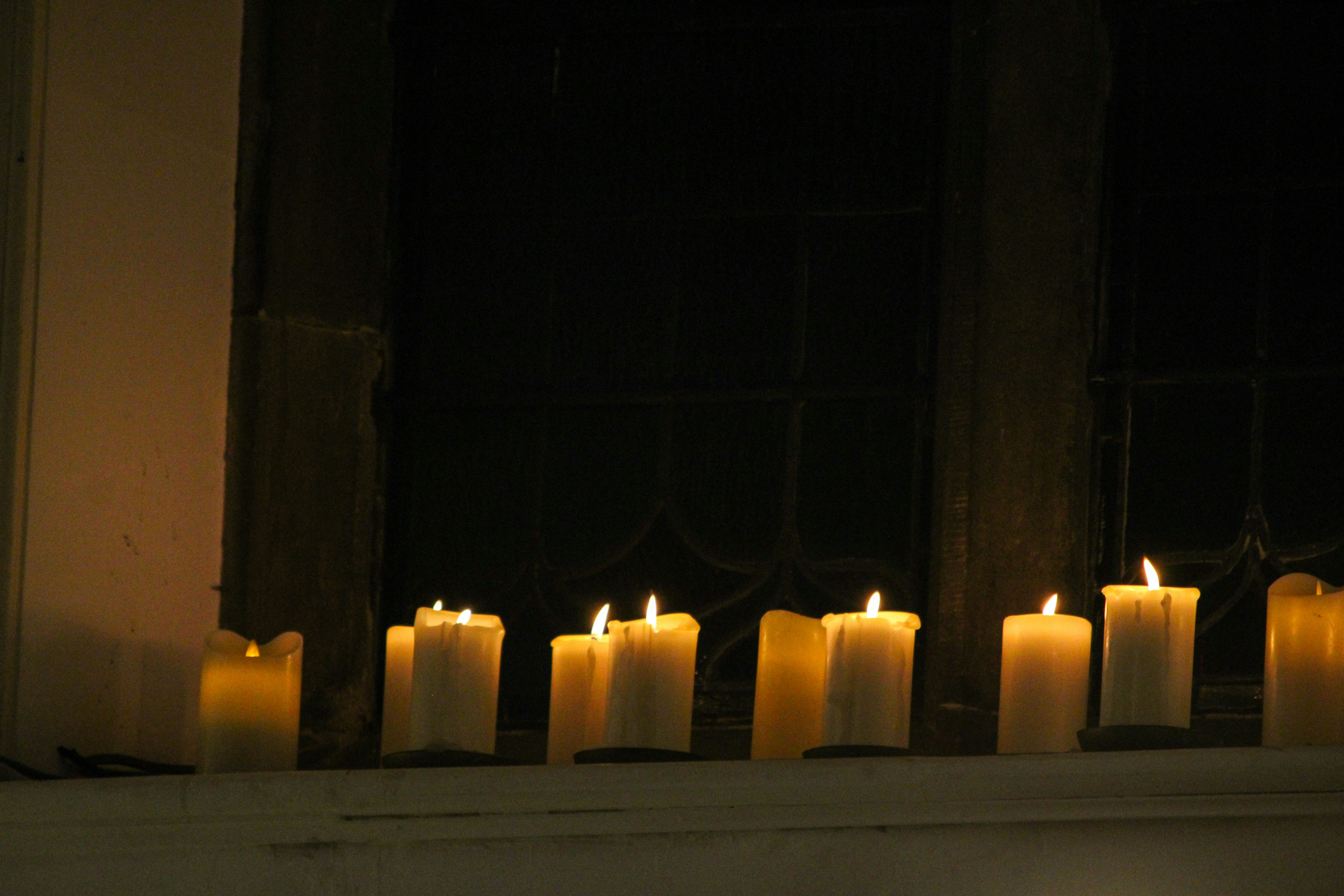 A row of lit white candles on a ledge before a dark window