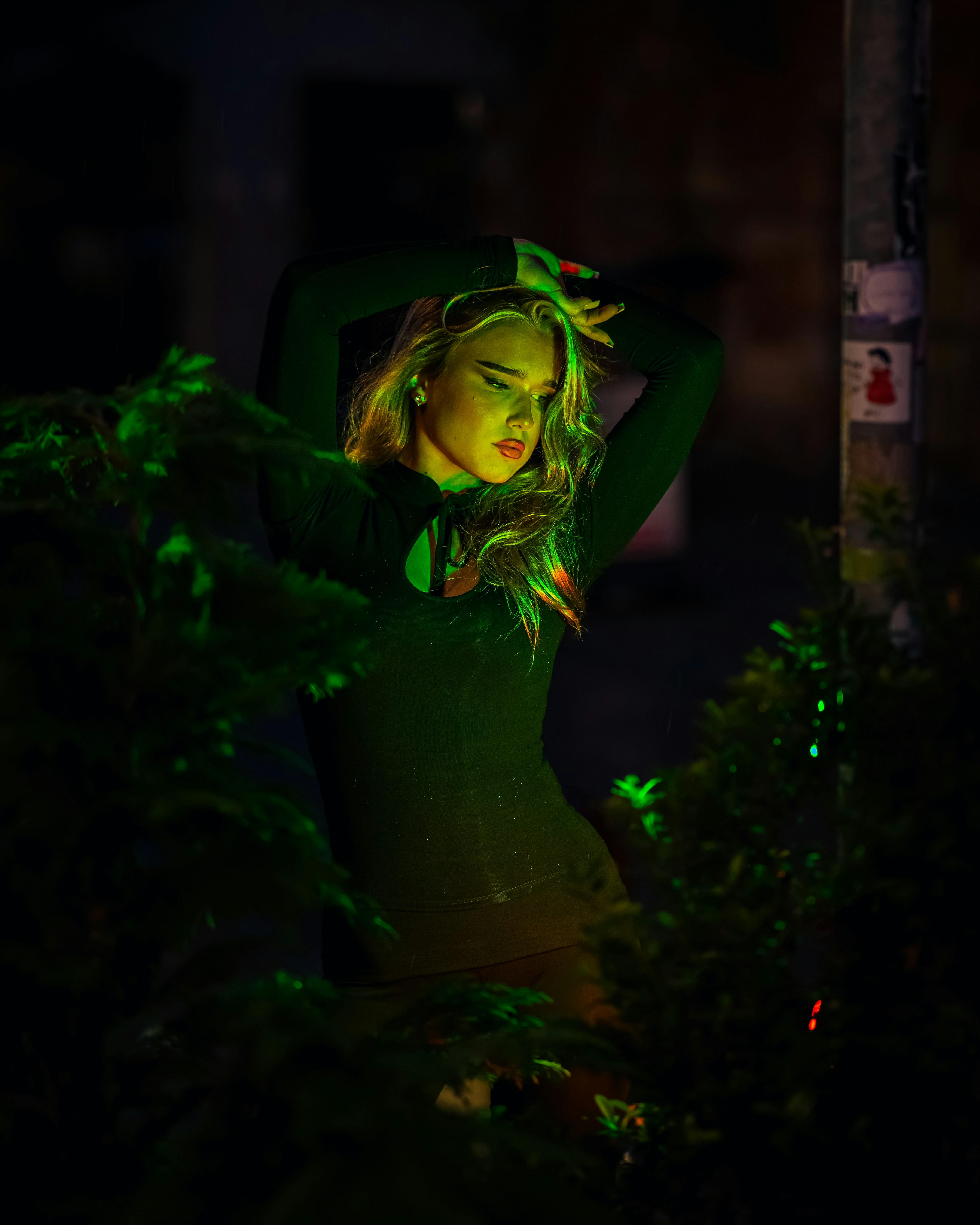 A woman in a black top lit by green light among dark foliage at night