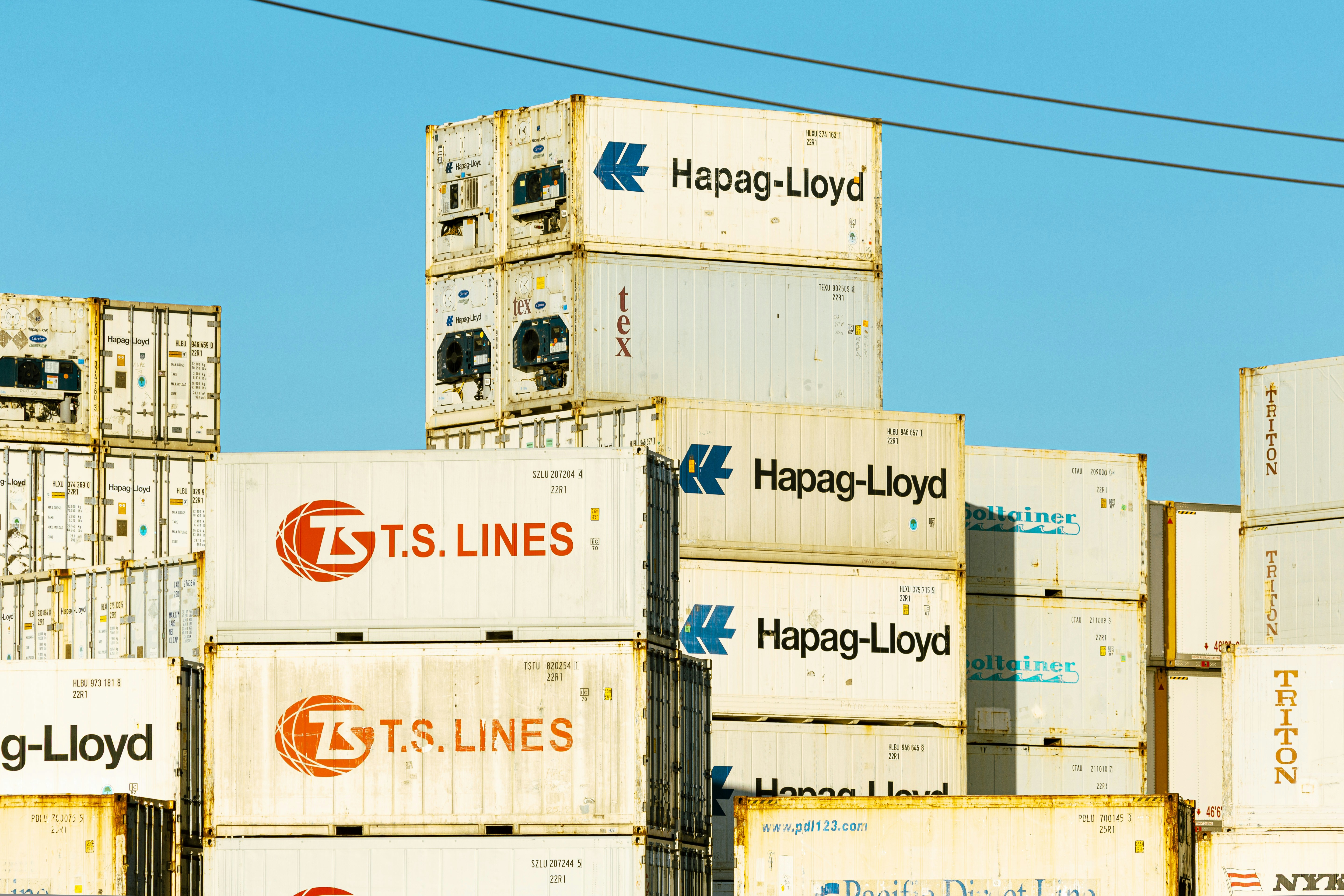 Stacked shipping containers with Hapag-Lloyd and T.S. Lines logos against a clear blue sky