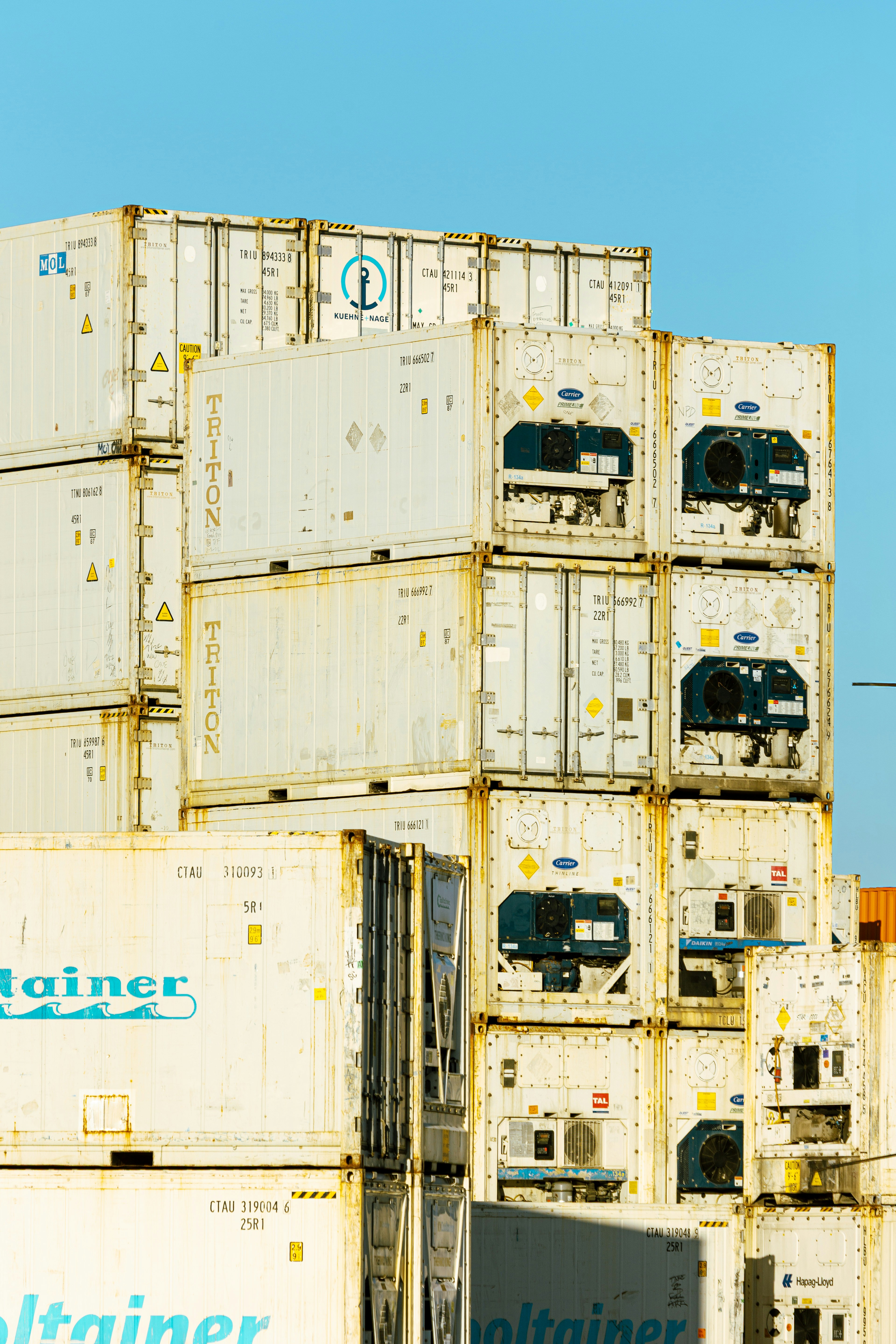 Stacked white shipping containers with refrigeration units against a clear blue sky