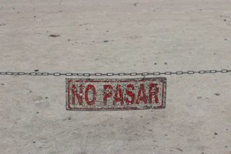 A weathered sign reading "NO PASAR" hanging from a metal chain over sand