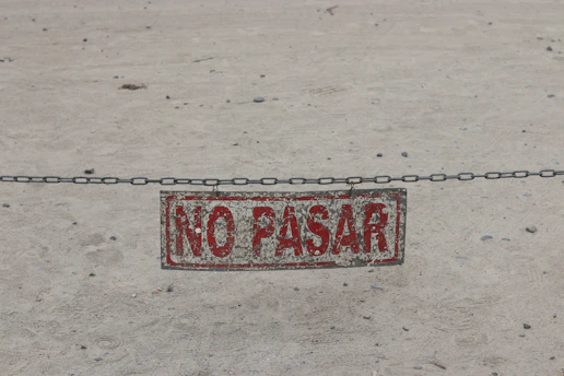 A weathered sign reading "NO PASAR" hanging from a metal chain over sand