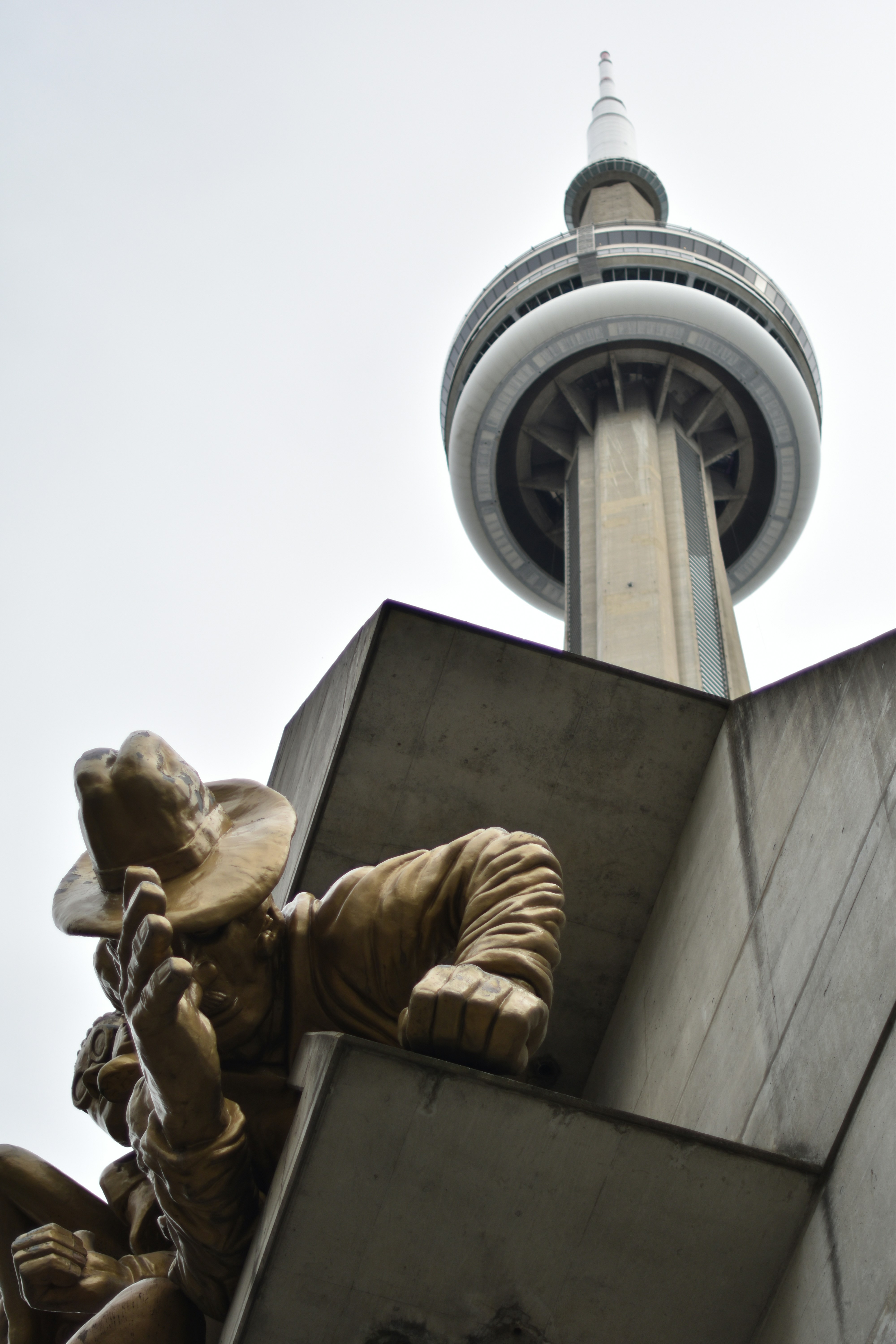 A golden statue of a person in a hat below the CN Tower in Toronto