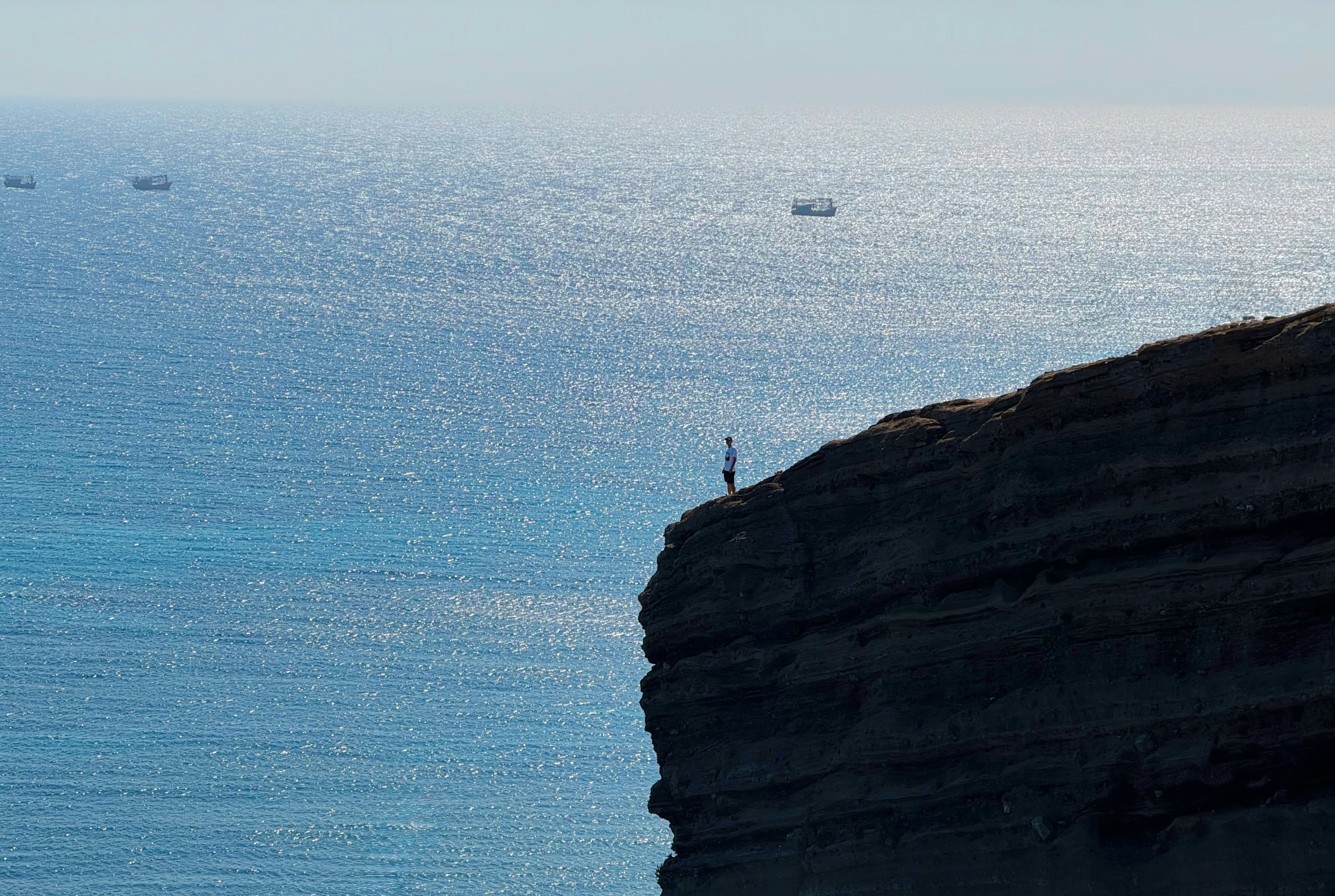 A person on a high rocky cliff overlooking the sparkling blue ocean
