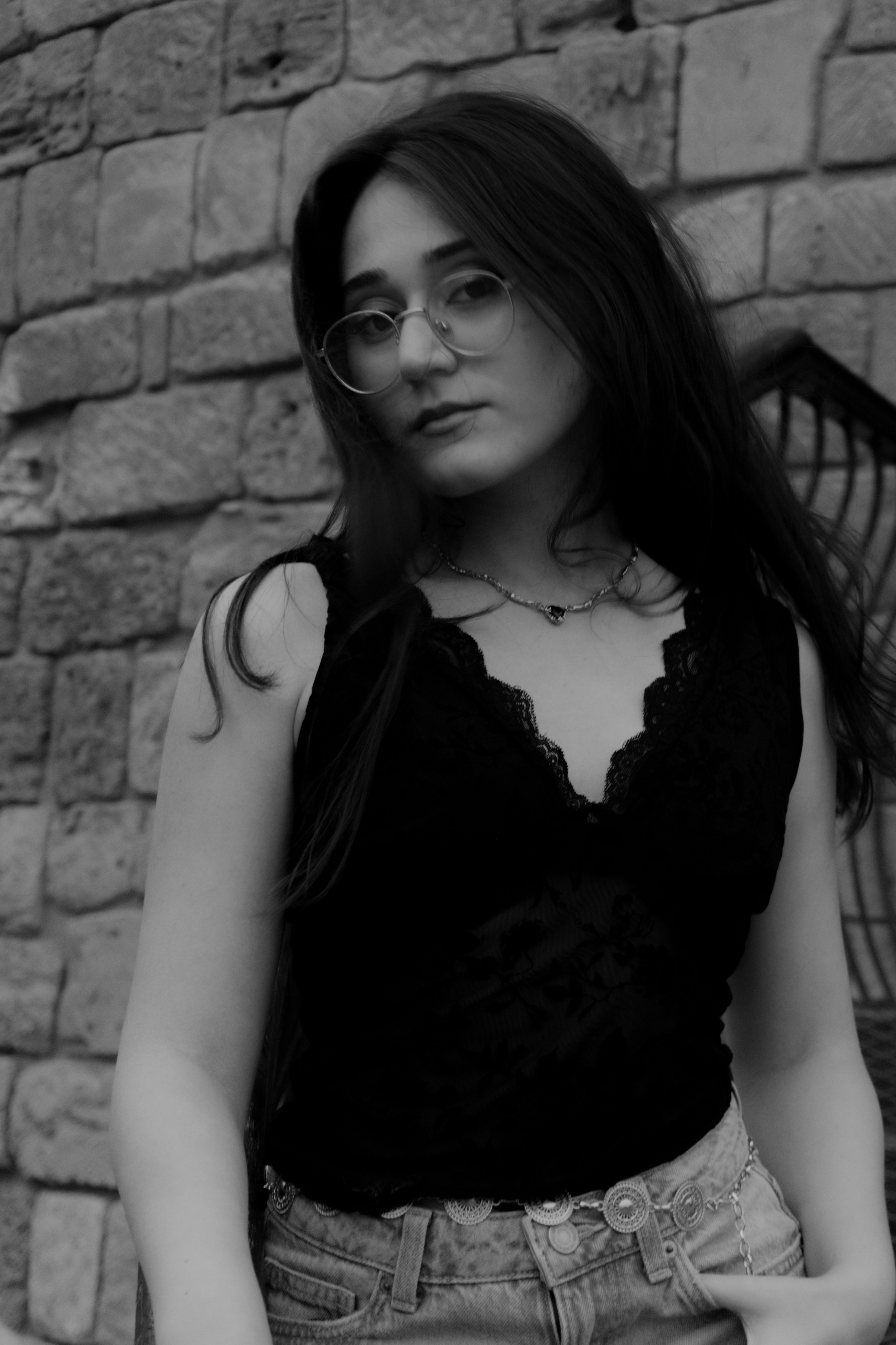 A young woman with glasses wearing a black lace top against a stone wall