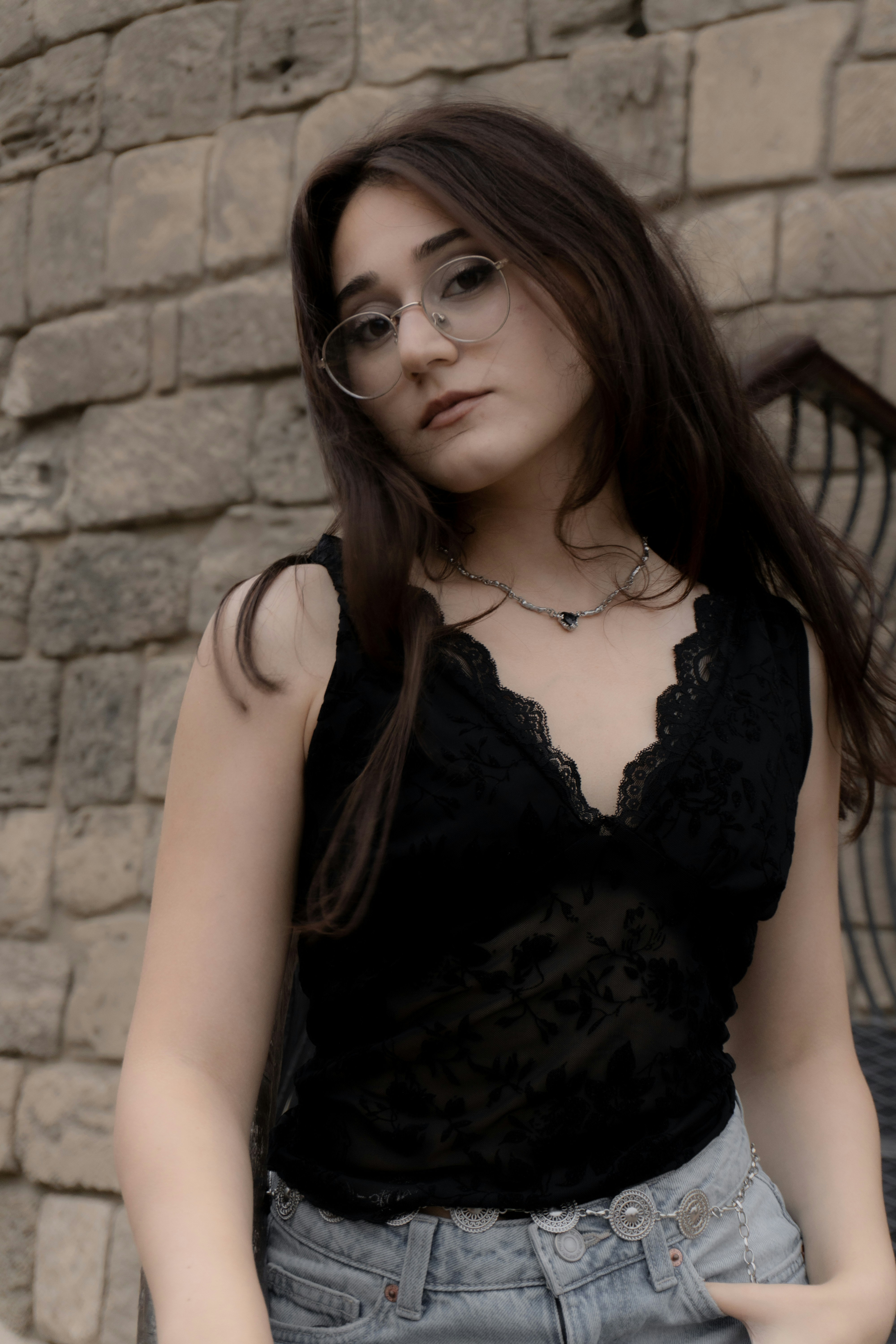 A woman with glasses and a black lace top against a stone wall