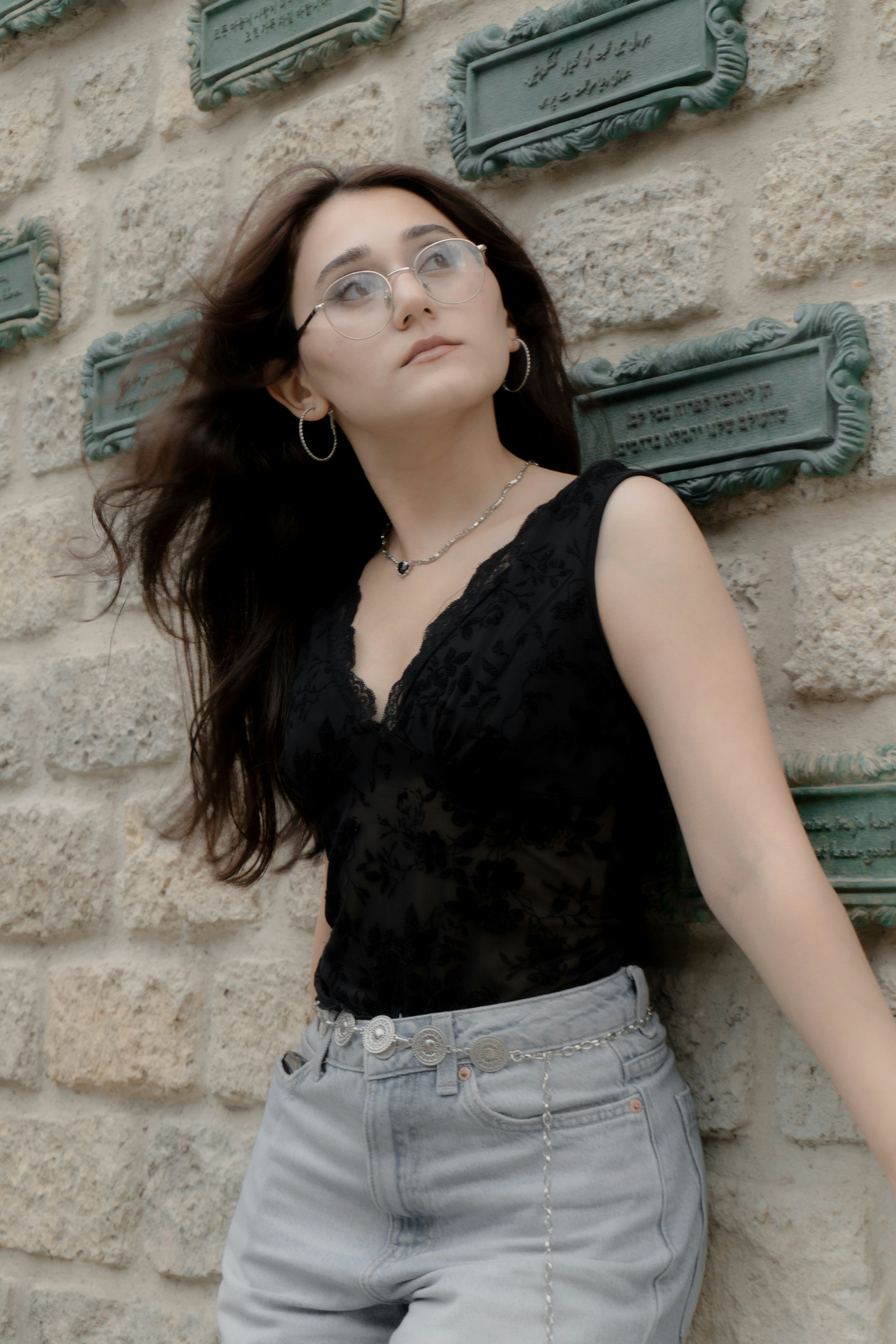 A woman in a black top and glasses leaning against a stone wall