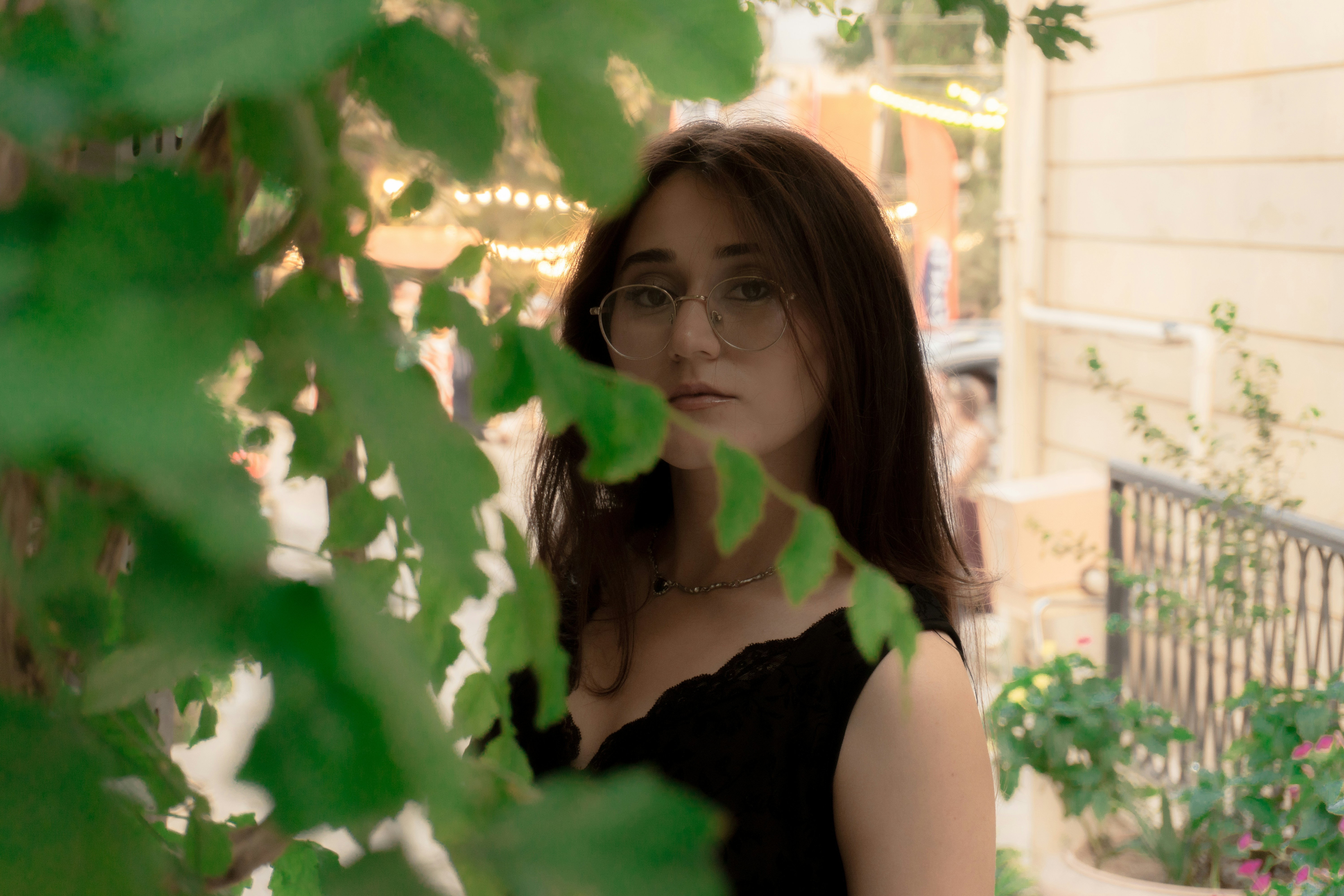 A woman with glasses and dark hair framed by green leaves outdoors