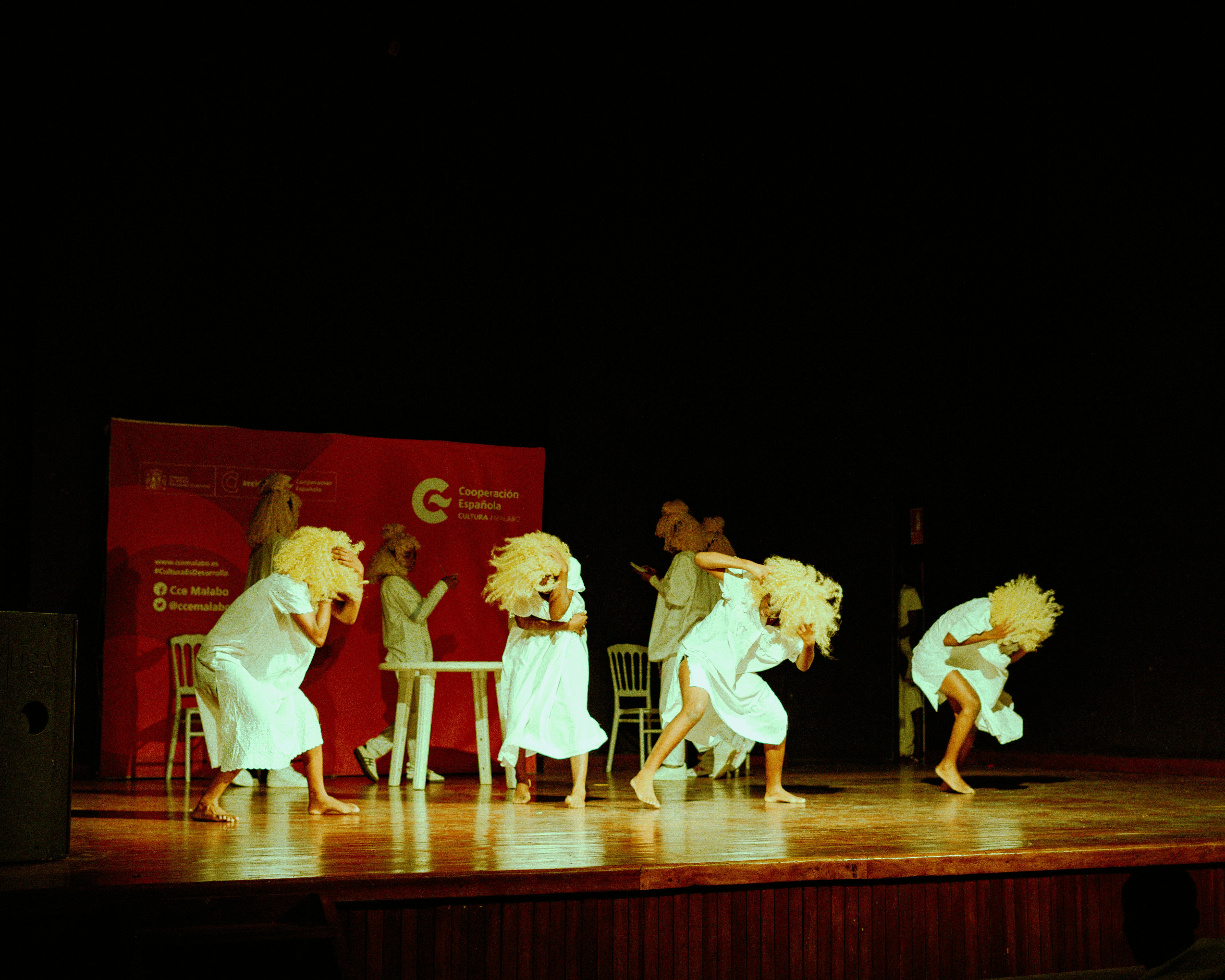 Performers in white dresses and blonde wigs dancing on a wooden stage