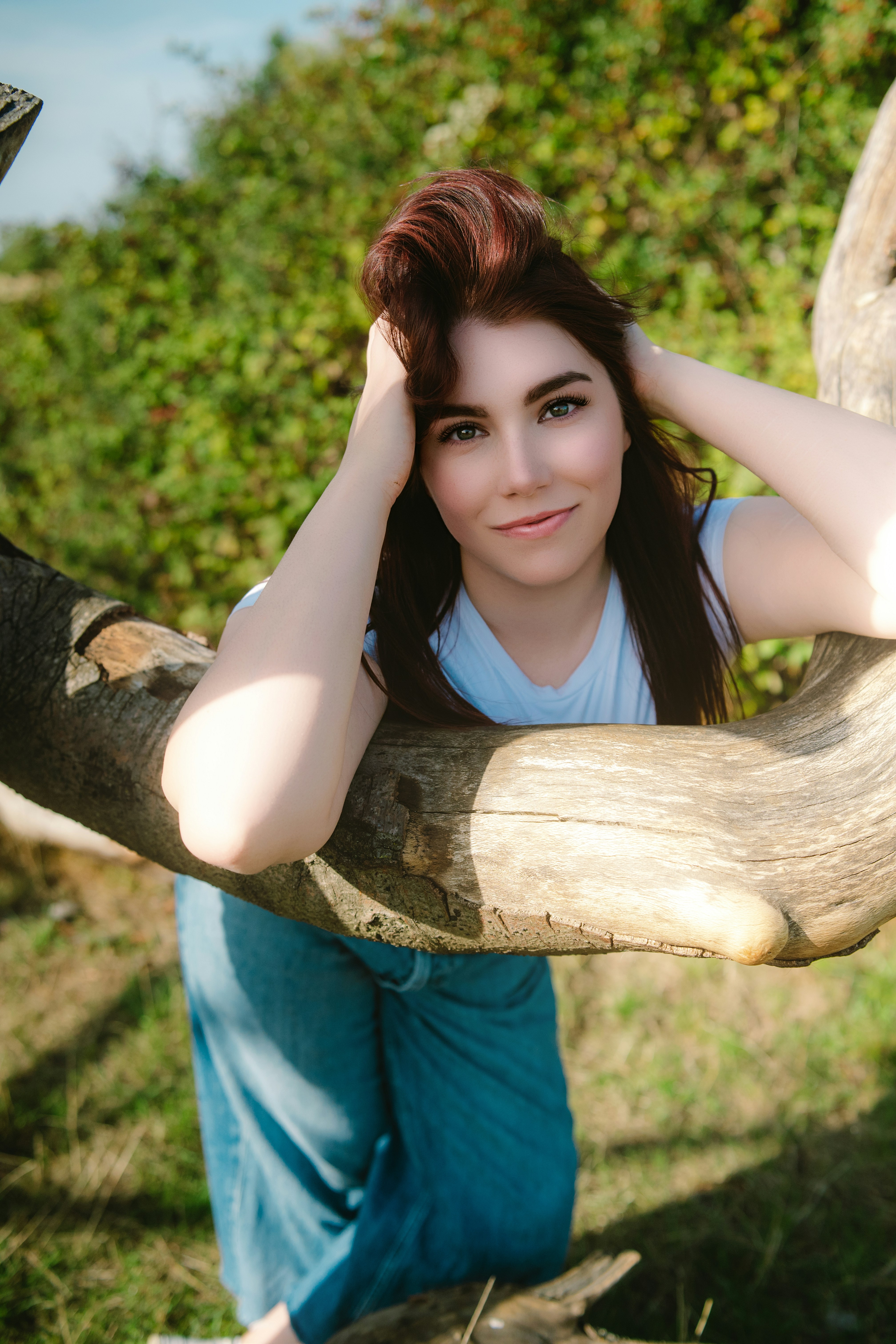 A woman with auburn hair leaning on a large tree branch outdoors