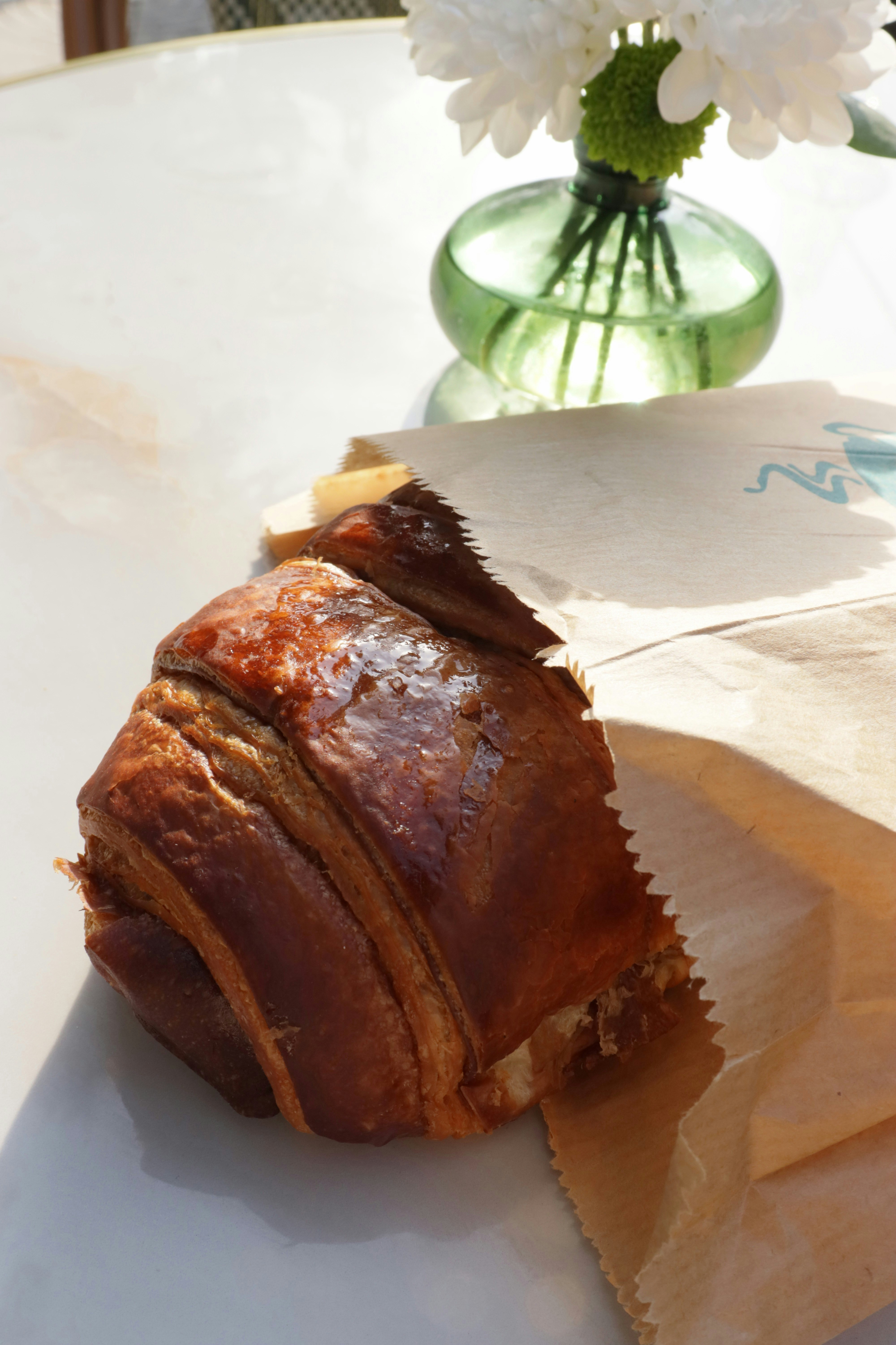 A glazed chocolate croissant in a paper bag beside a small green vase