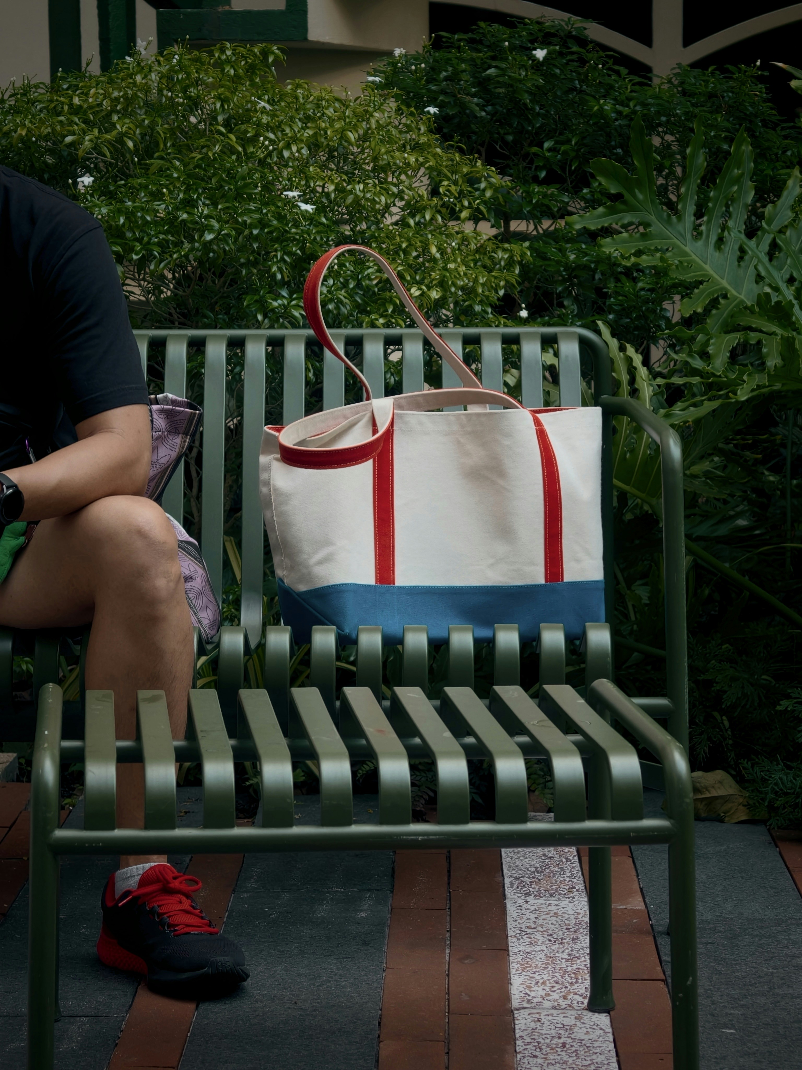 A person sitting on a green metal bench beside a canvas tote bag