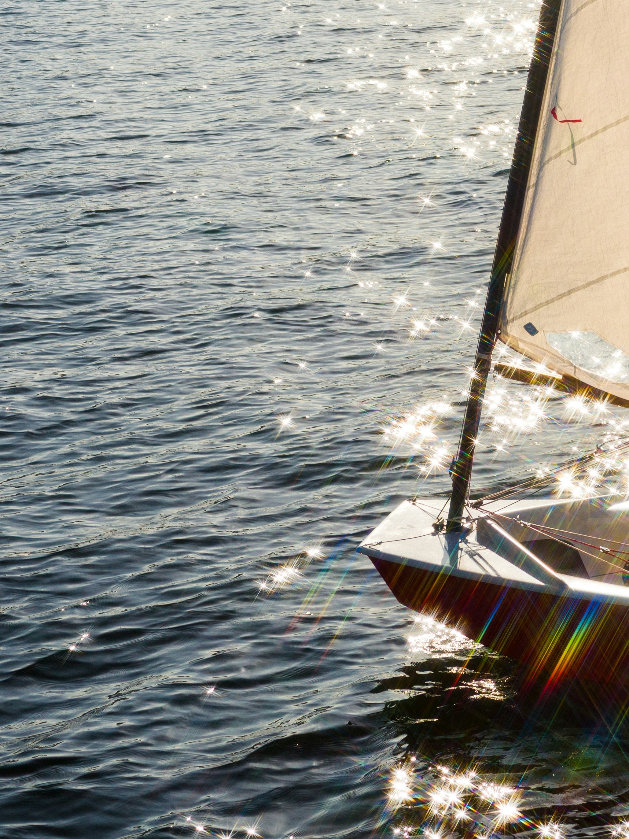 The bow of a sailboat on dark water with sparkling sunlight reflections