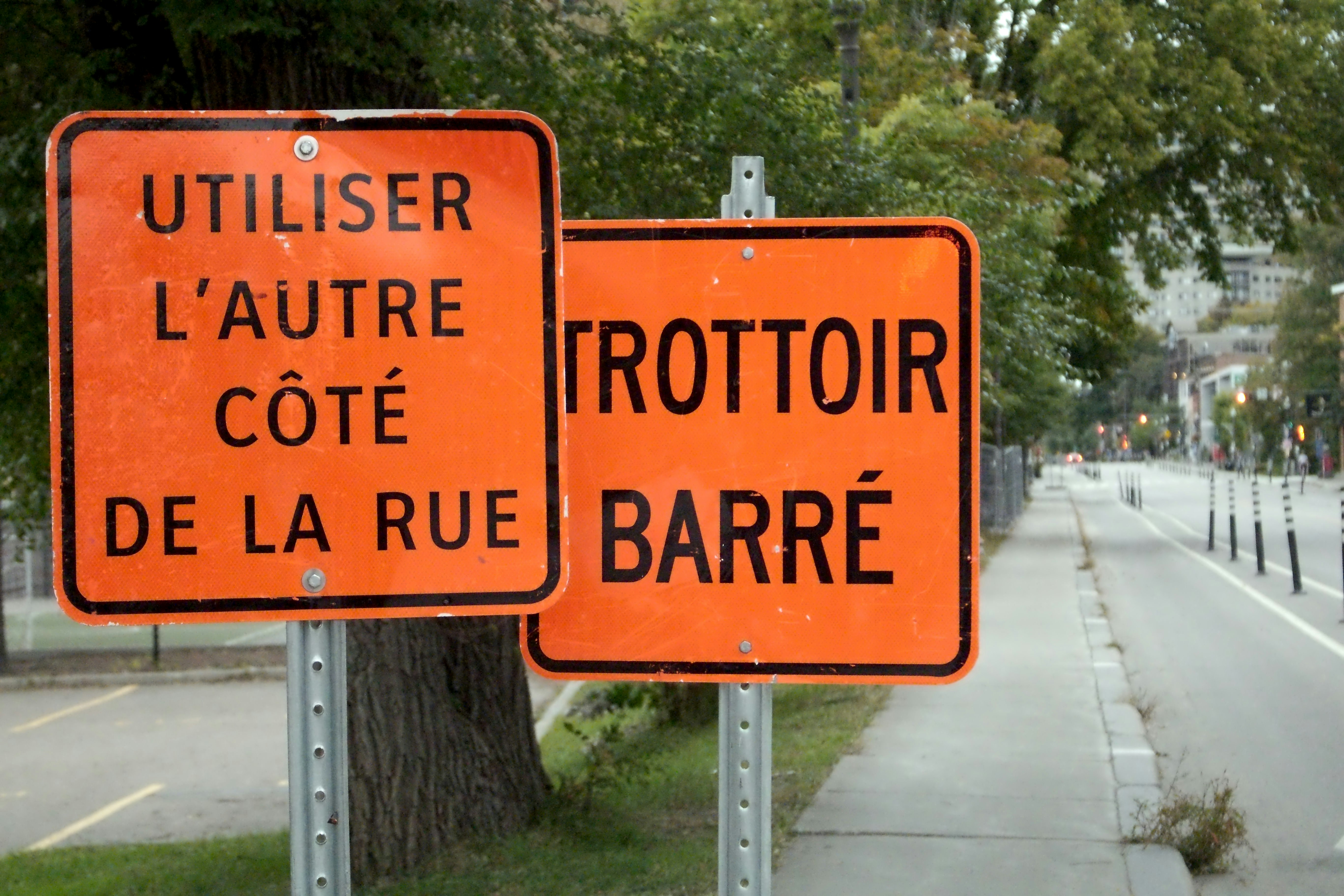 Two orange construction signs reading "UTILISER L'AUTRE CÔTÉ DE LA RUE" and "TROTTOIR BARRÉ"