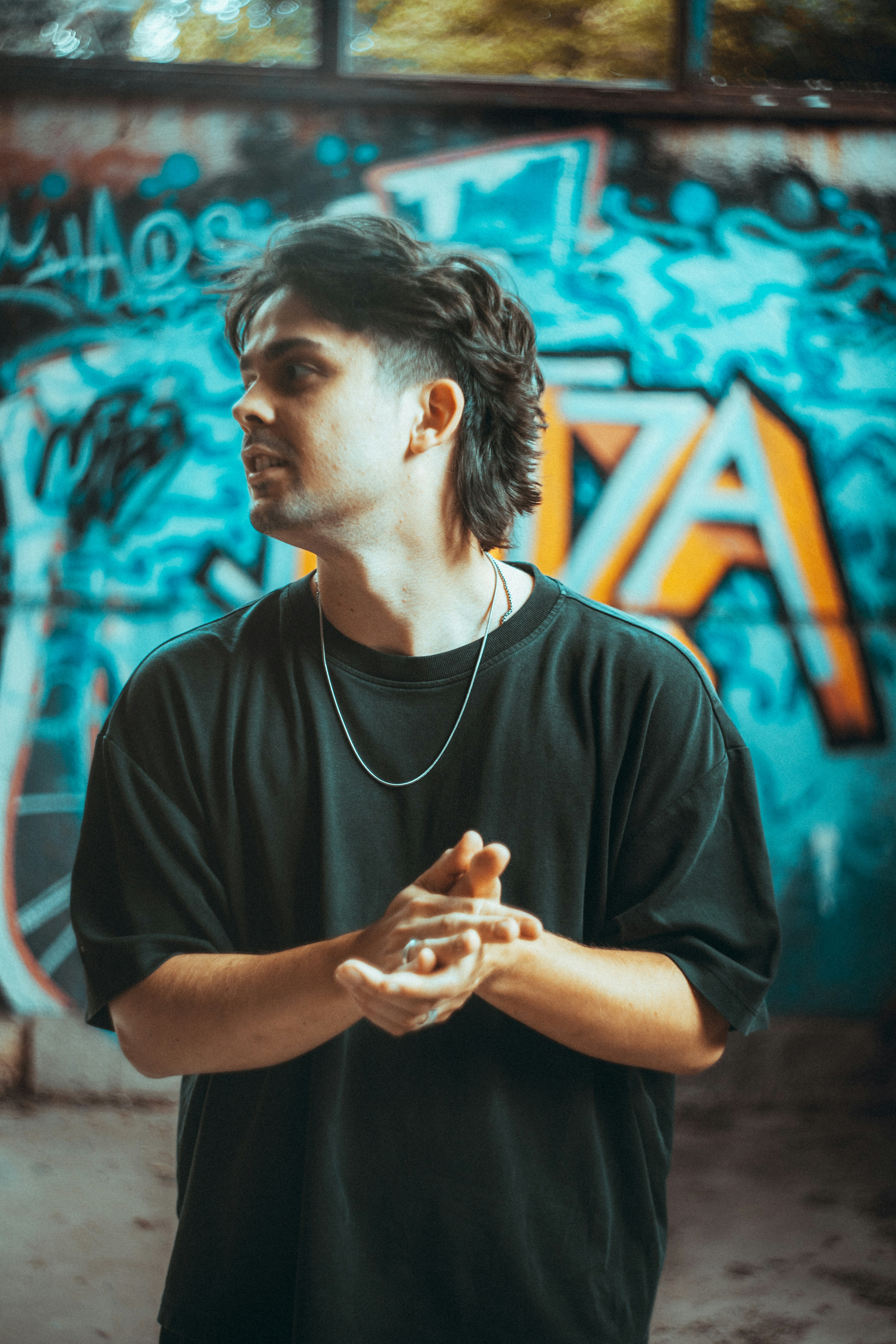 A man with a mullet wearing a black t-shirt and silver chain against graffiti