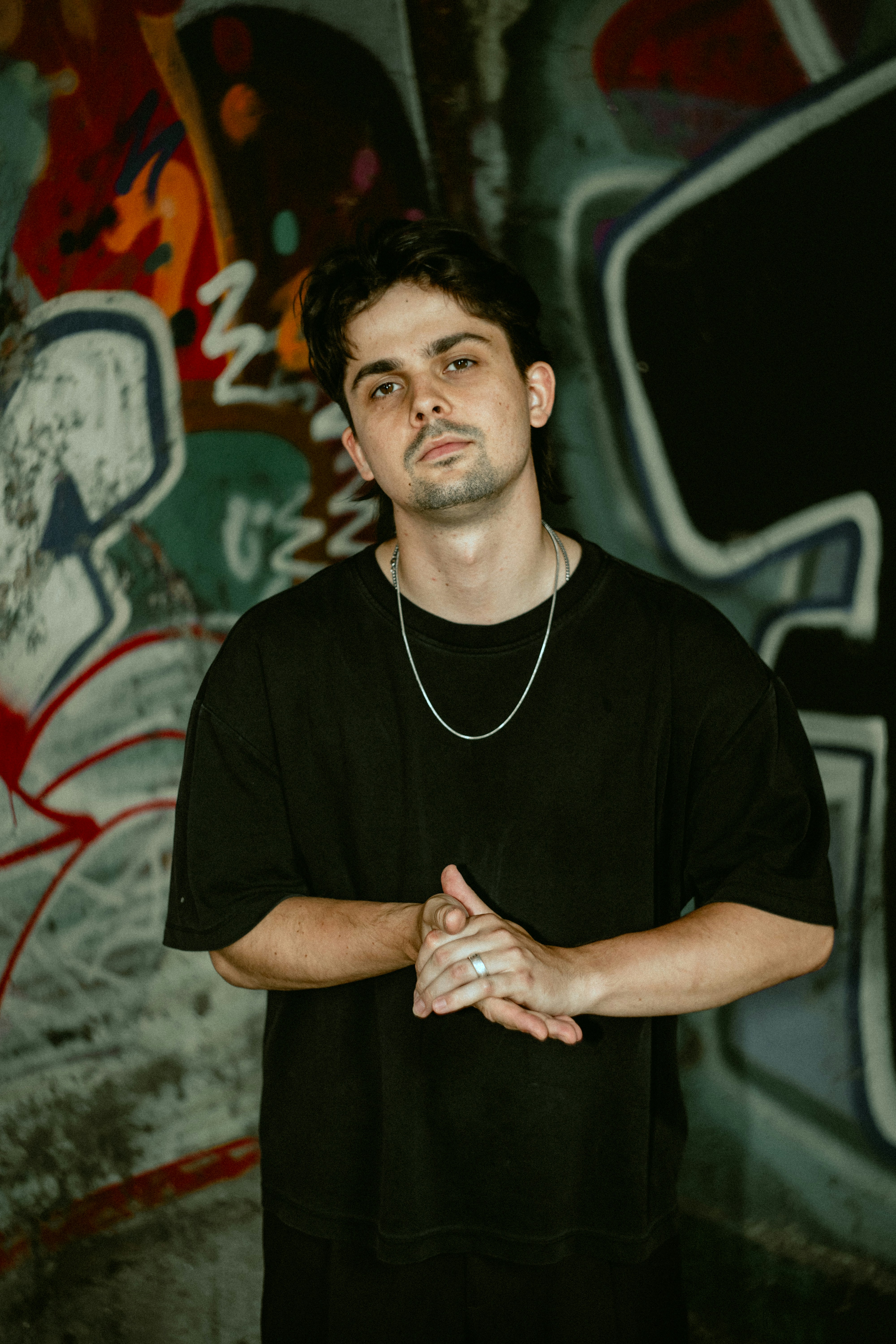 A man in a black t-shirt and silver chain against a graffiti wall