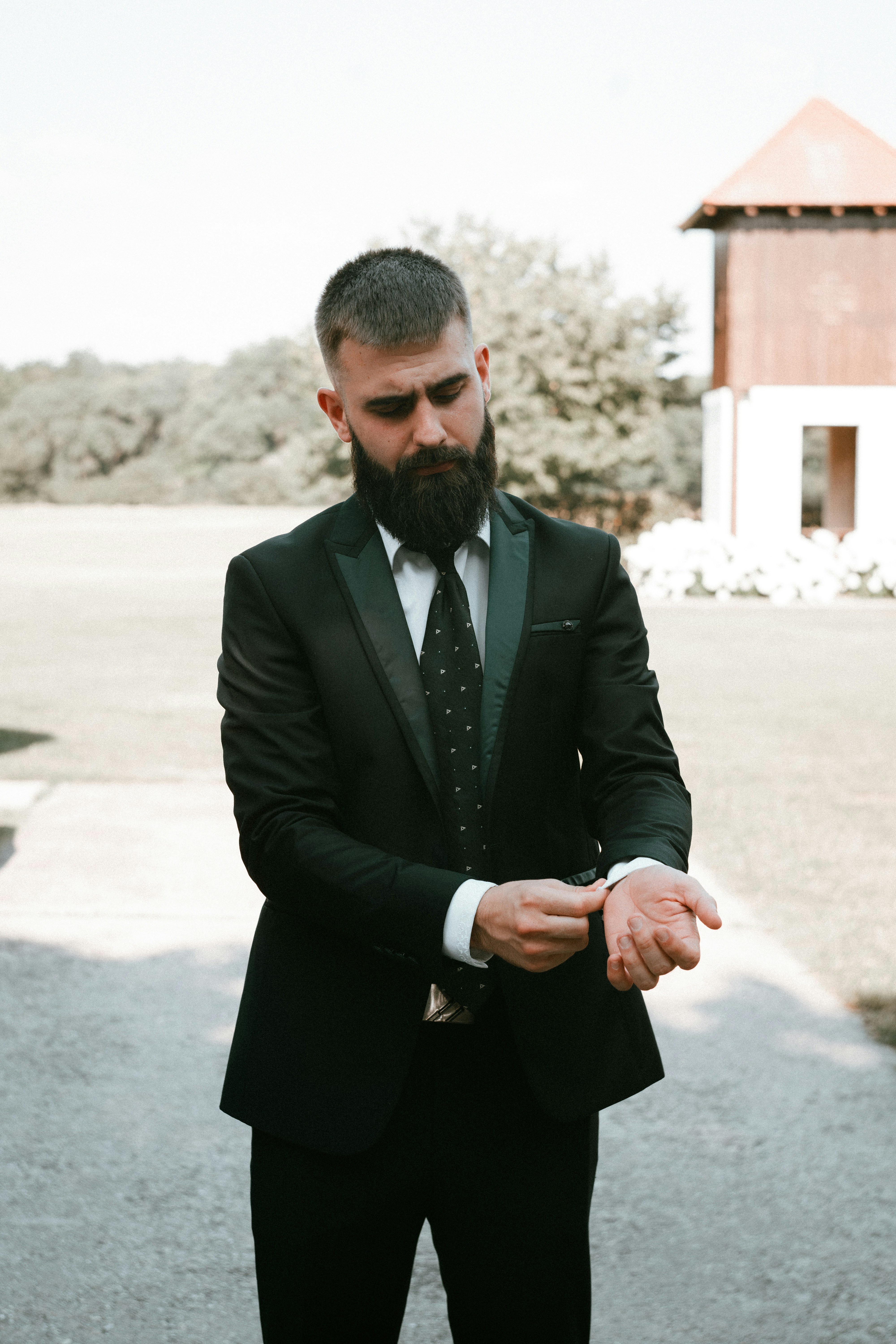 A man with a beard in a black suit adjusting his white shirt cuff