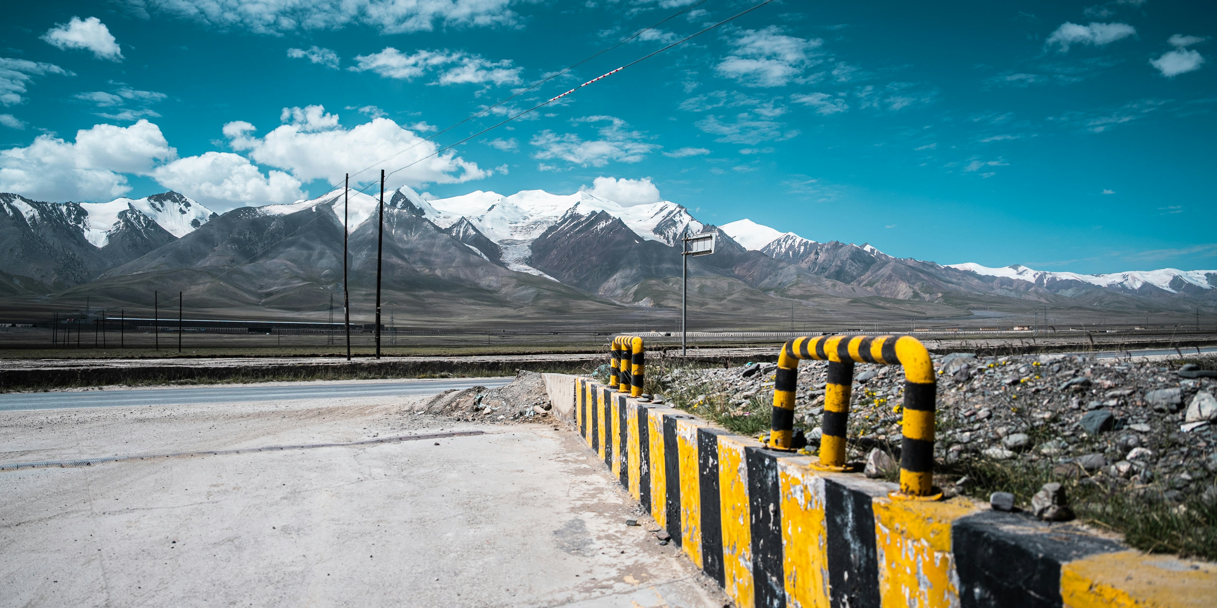 A road with black and yellow striped barriers before snow-capped mountains under a blue sky