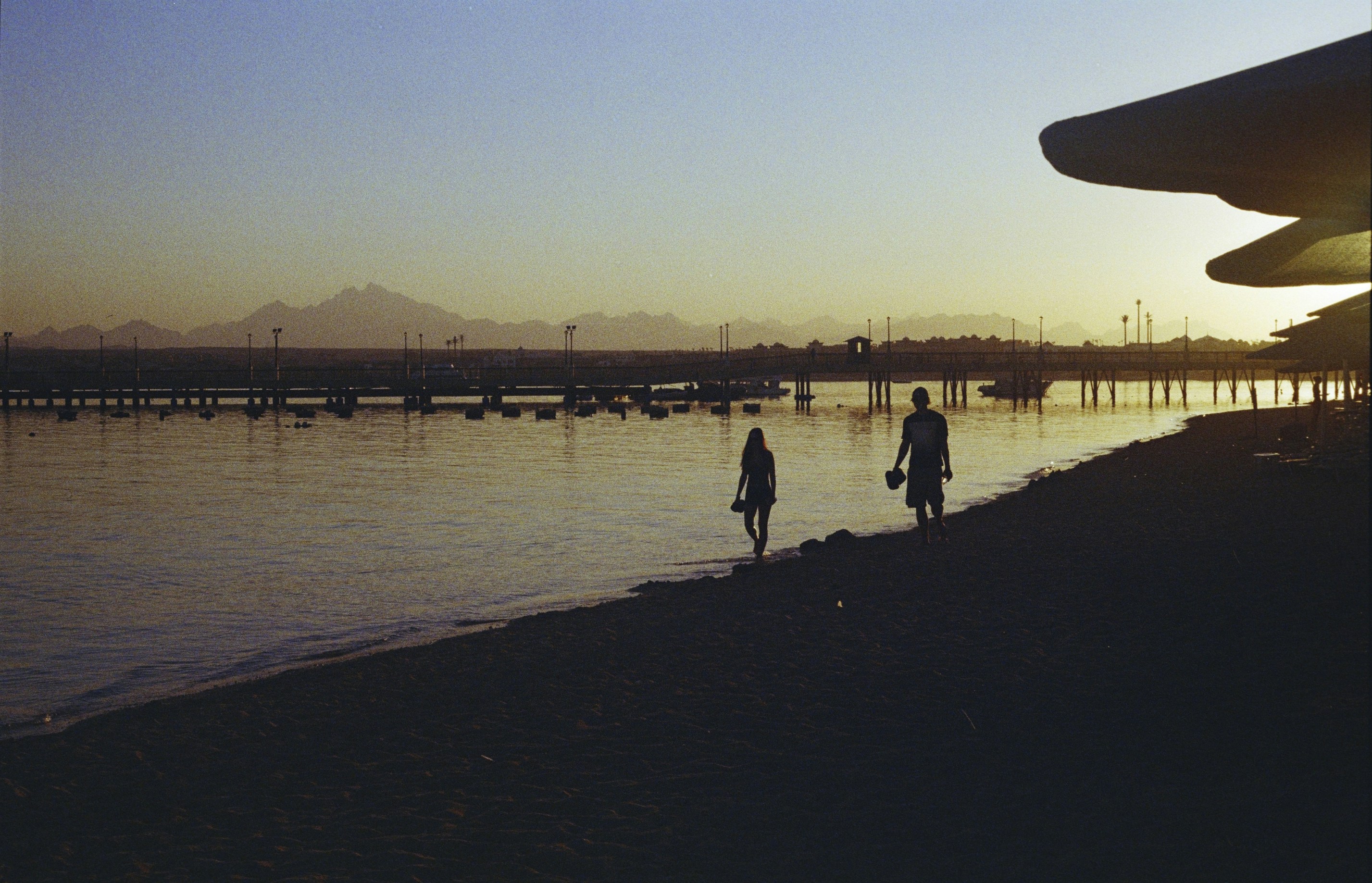 Two people in silhouette walking along a beach near a wooden pier at low sun