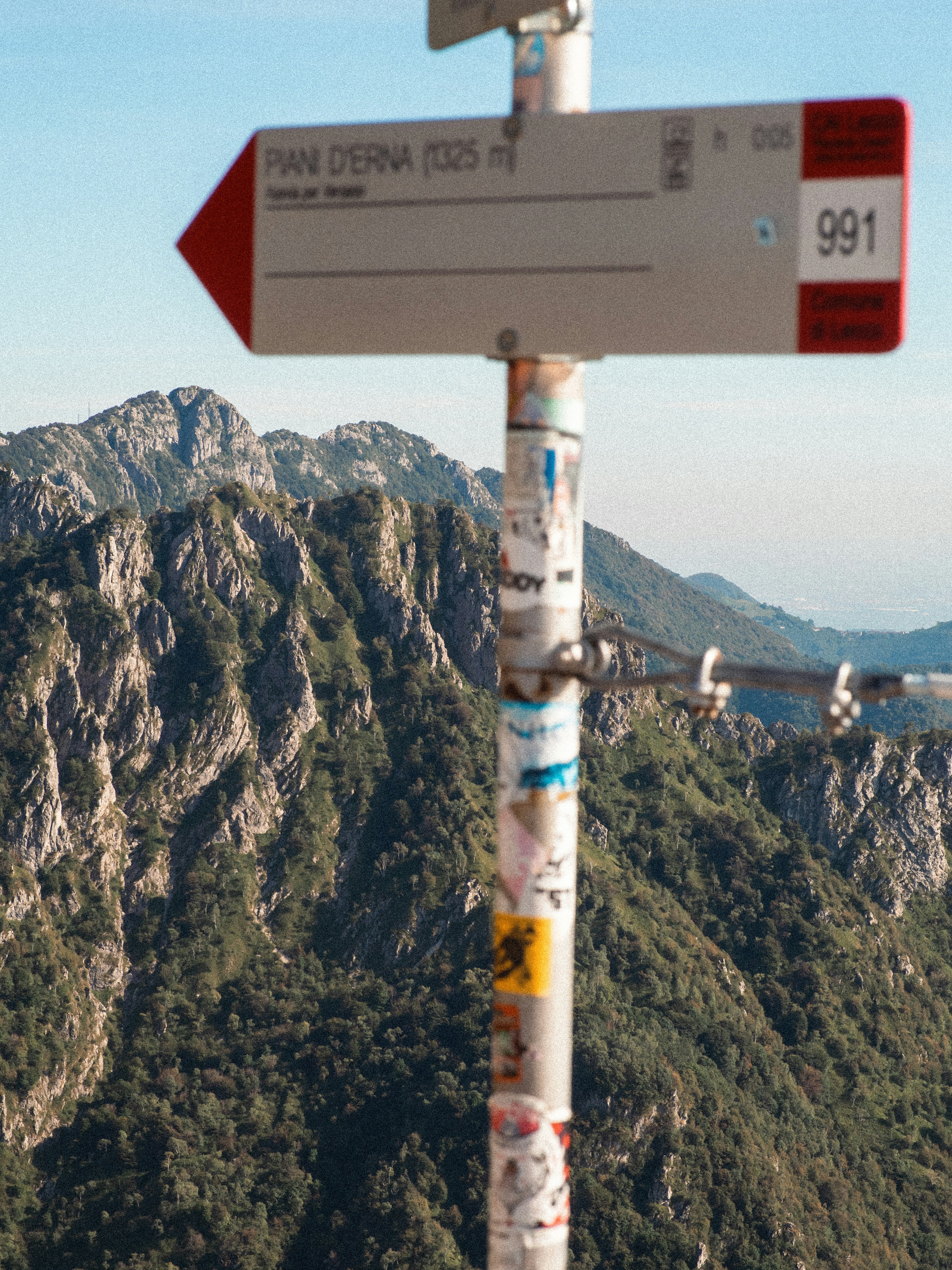 A hiking trail sign pointing toward Piani d'Erna against a backdrop of rocky mountains