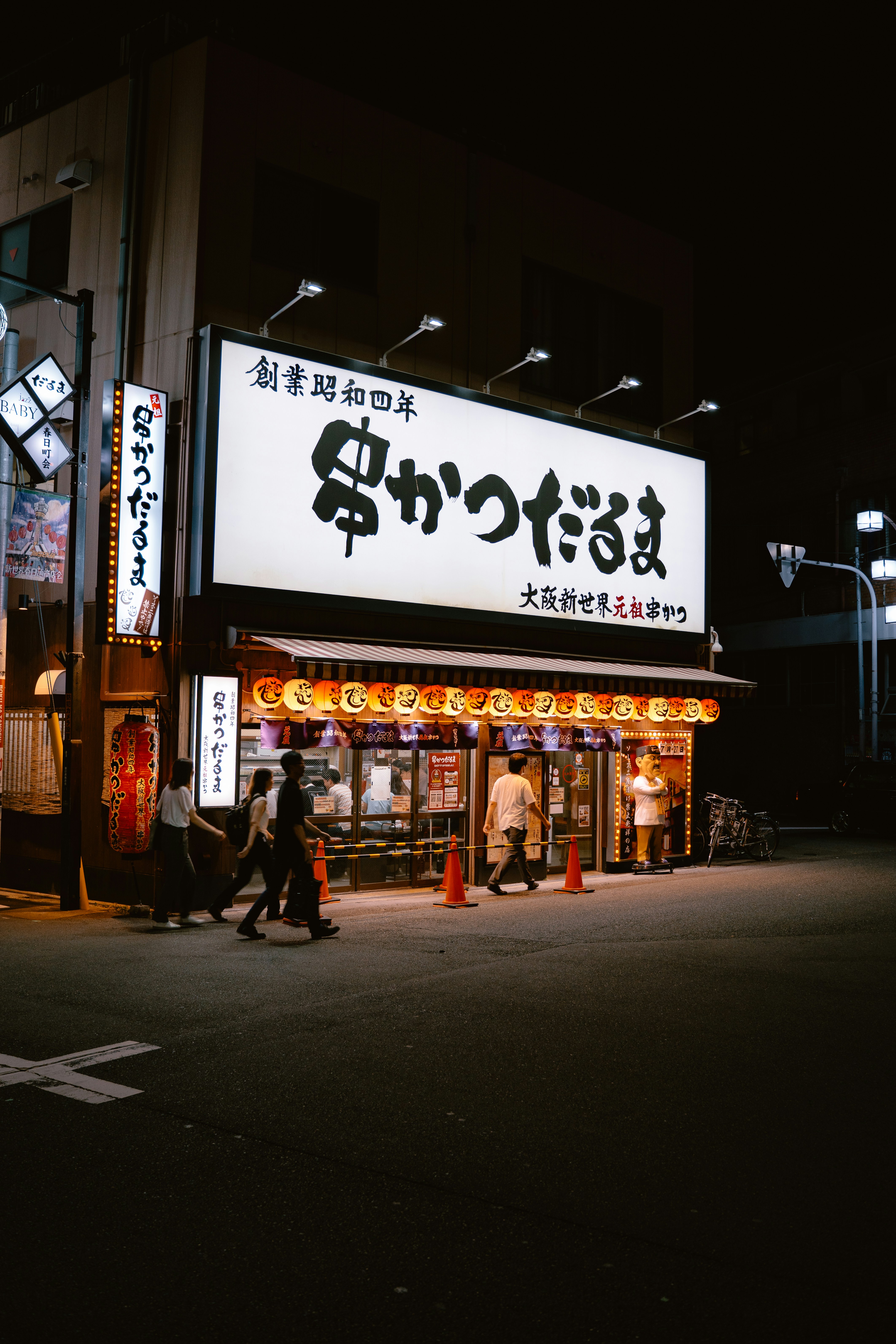 People walking past a brightly lit restaurant with paper lanterns in Osaka at night