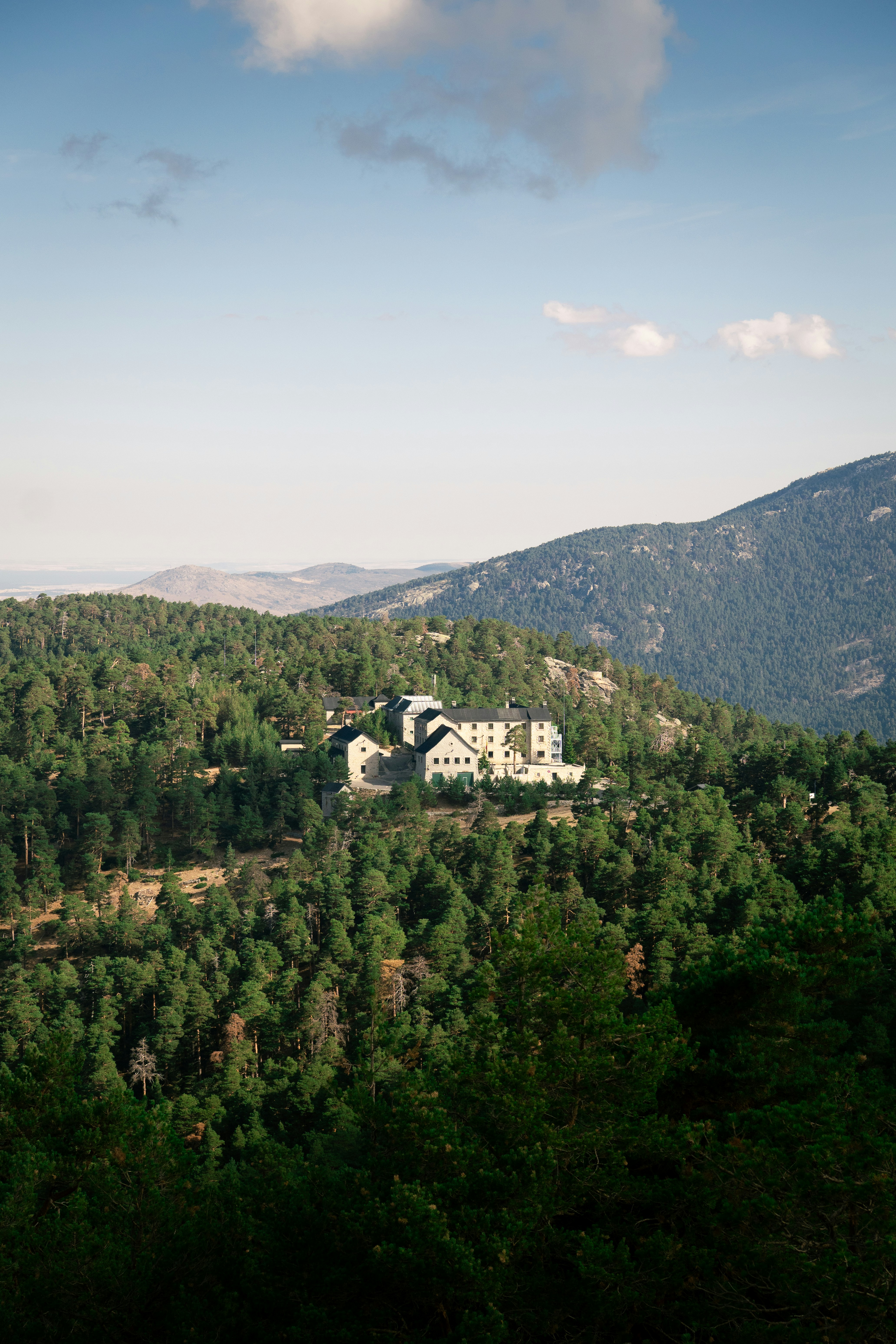 A stone building complex nestled within a dense pine forest on a mountain slope