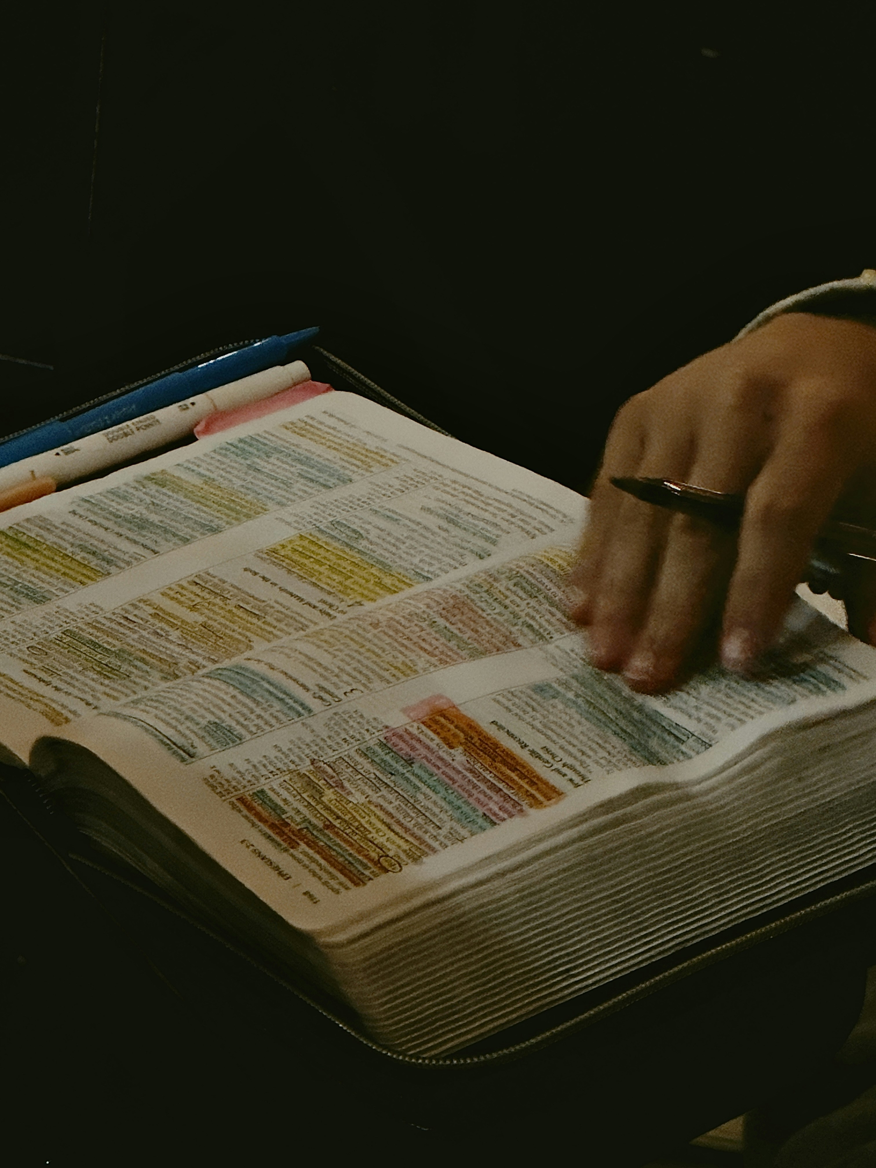 A hand resting on an open book with colorful highlighted text and pens