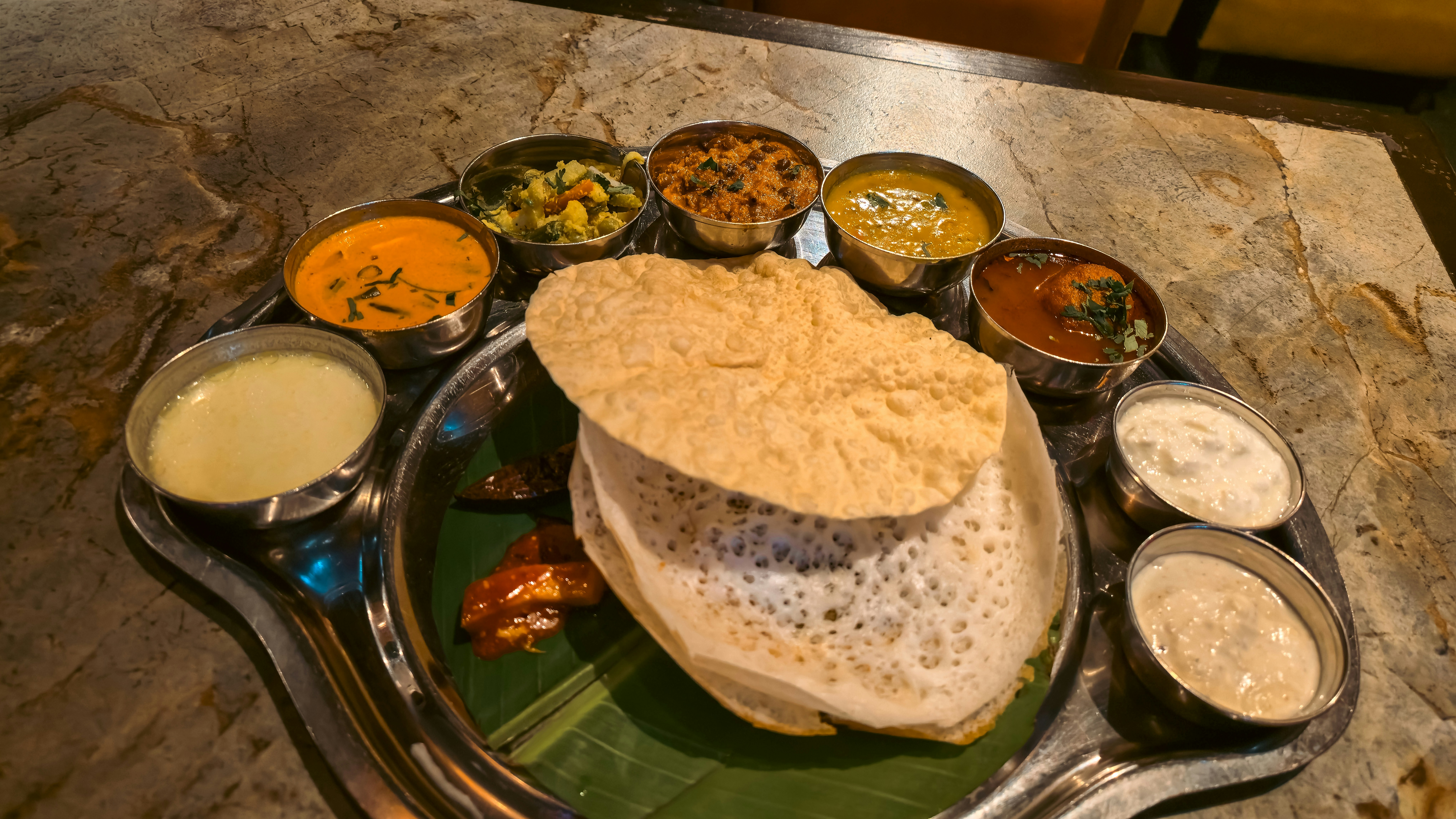 A South Indian thali on a banana leaf: appams and a pappadum ringed by small steel bowls of curries and chutneys