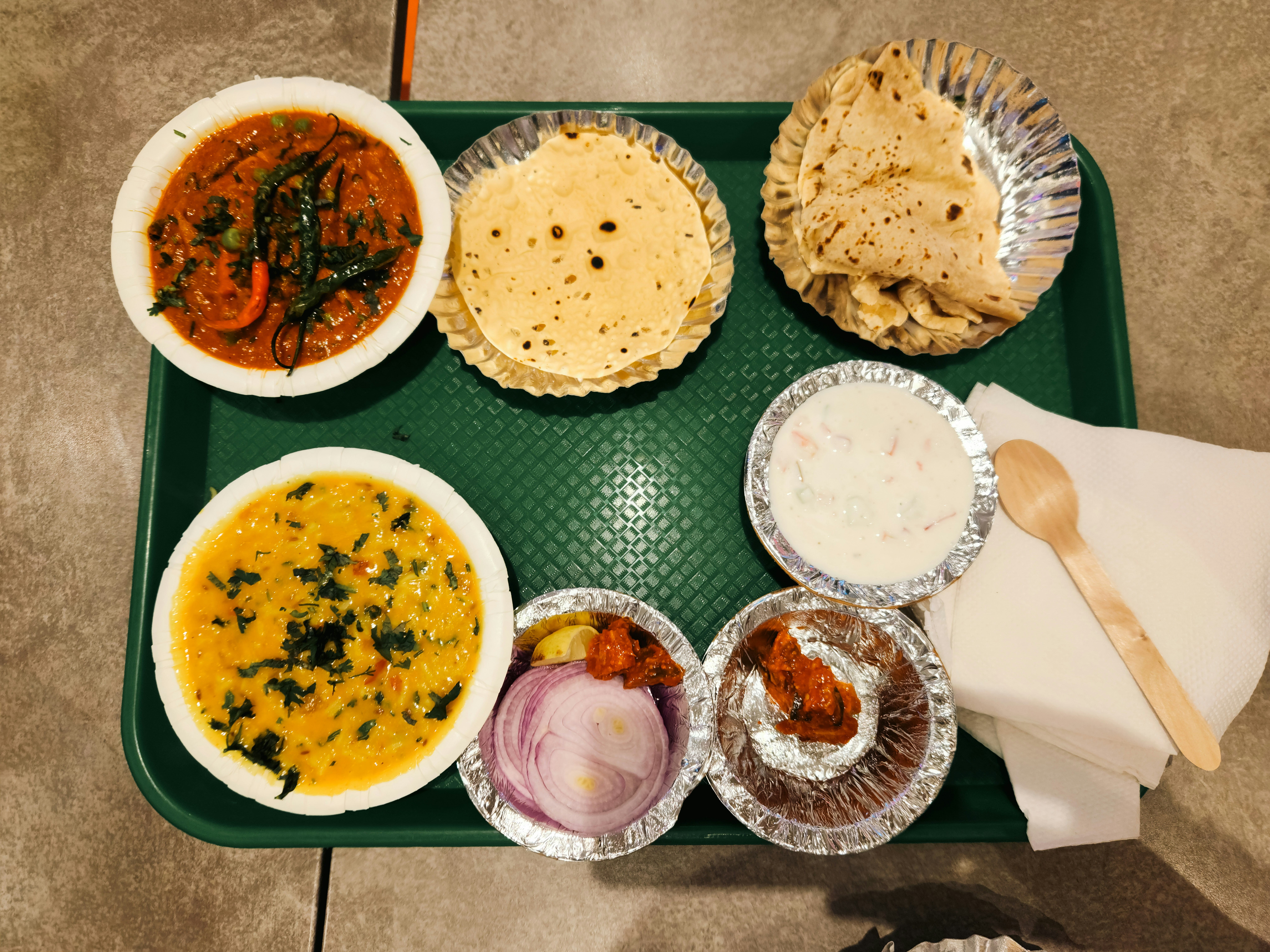 A green tray with Indian curry, dal, roti, raita, onions, and pickle