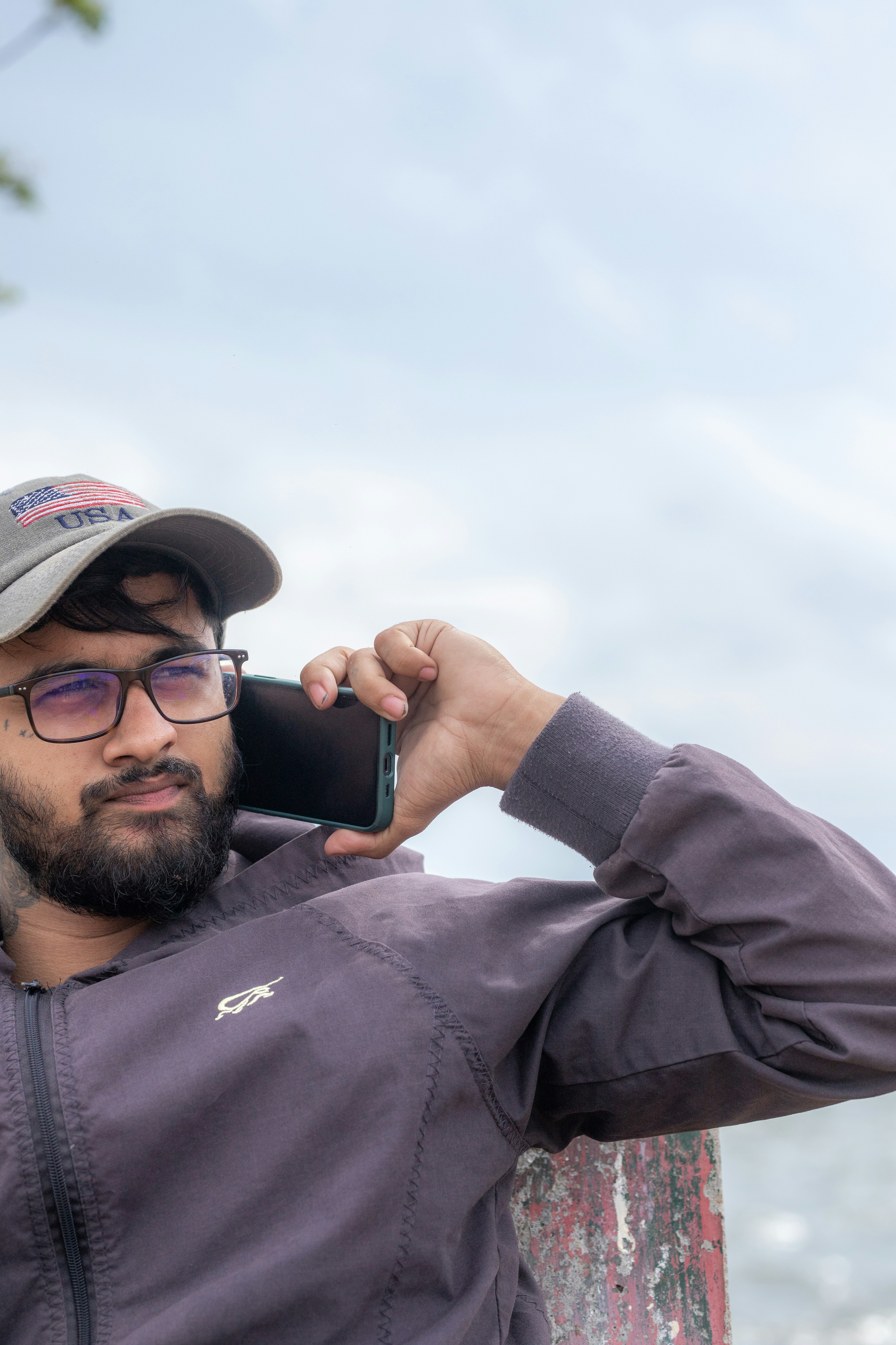 A man with a beard and glasses wearing a cap talking on a smartphone