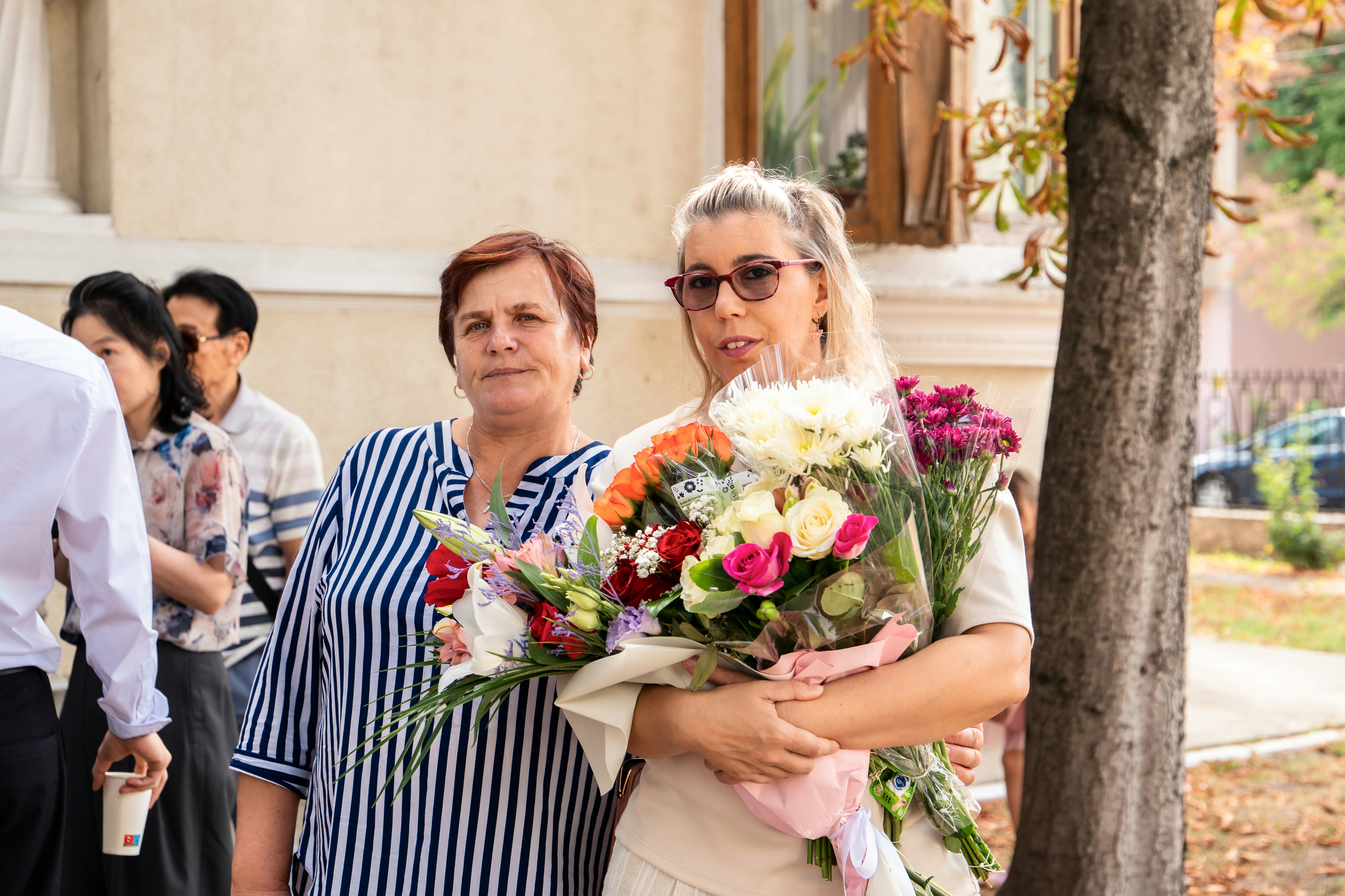 Two women standing together, one holding a large bouquet of colorful flowers outdoors