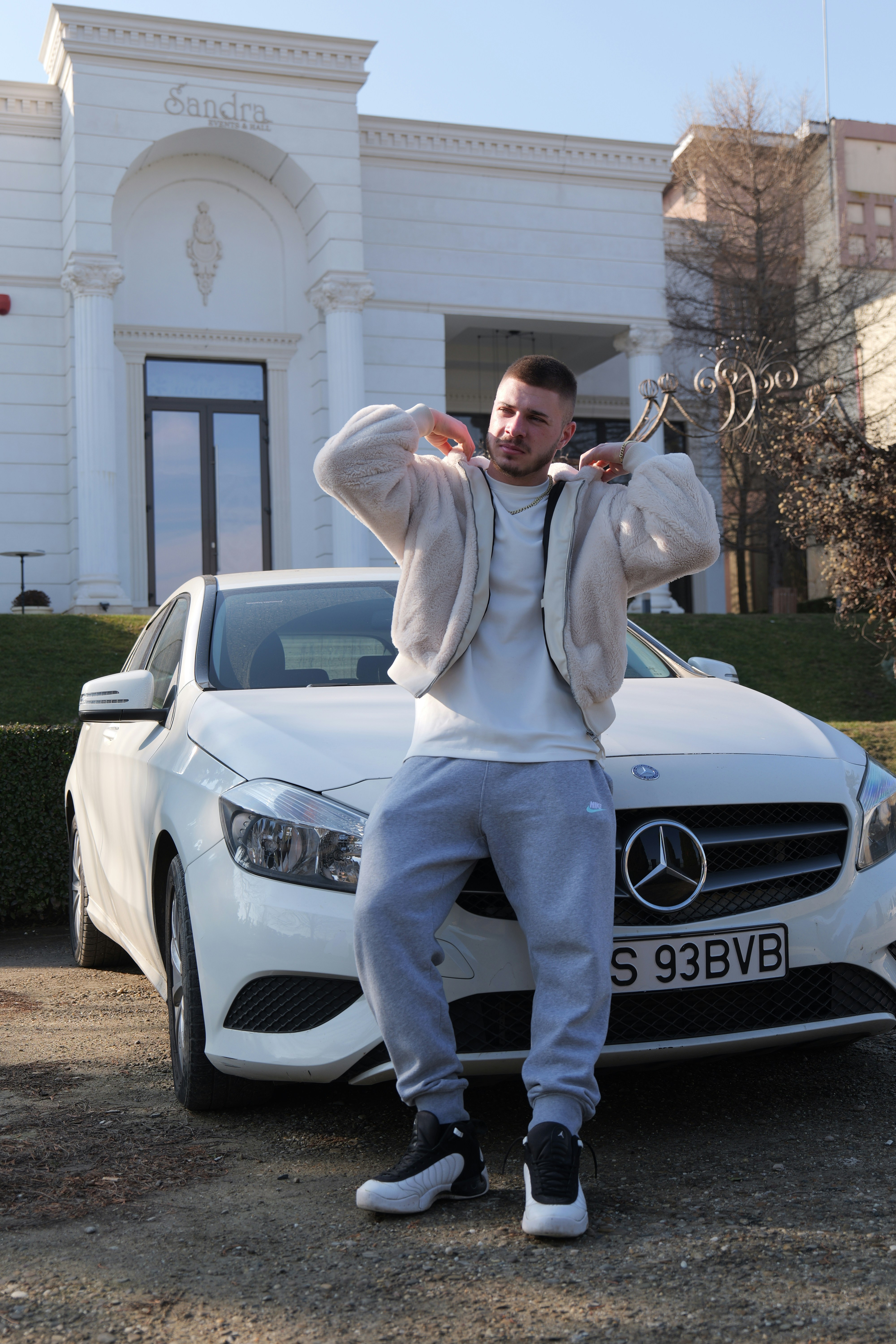 A man in a cream fleece jacket leaning against a white Mercedes car